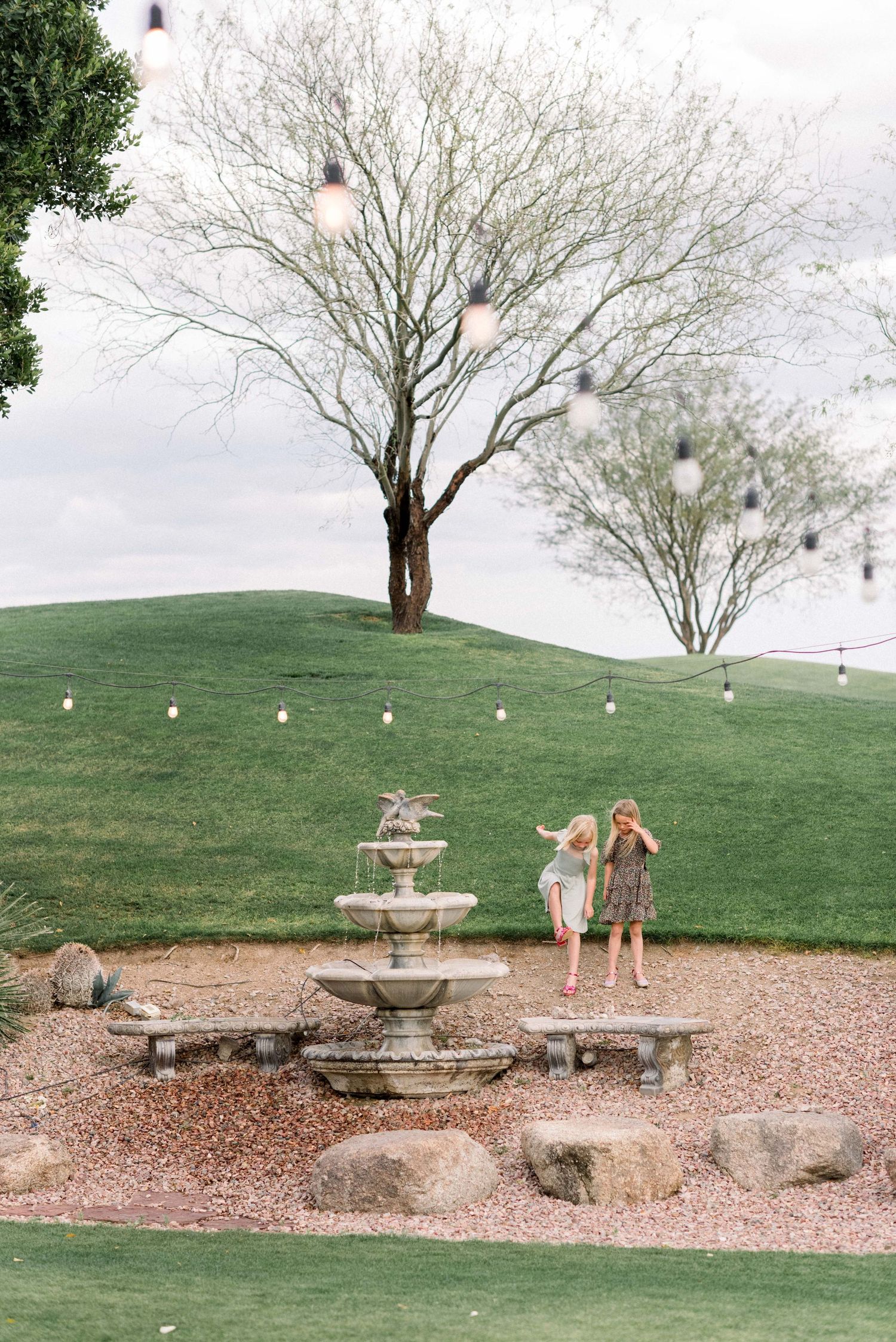 Scenic outdoor wedding venue with a decorative fountain and string lights on a grassy hill at dusk.
