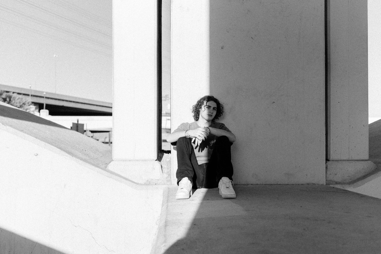 Black and white photo of person sitting against concrete wall in urban setting.