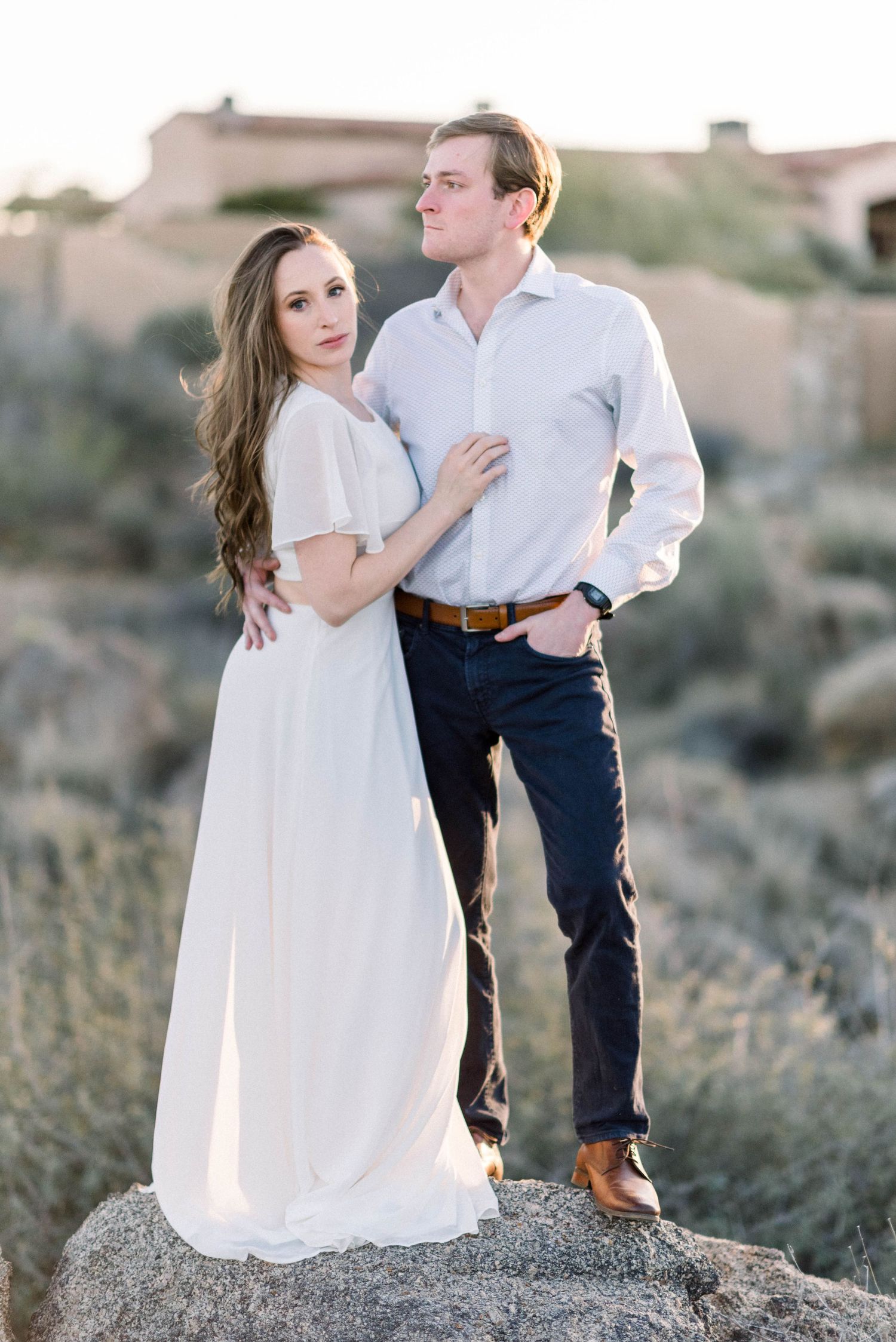 Desert engagement portrait on rocky terrain with flowing white dress and casual attire.