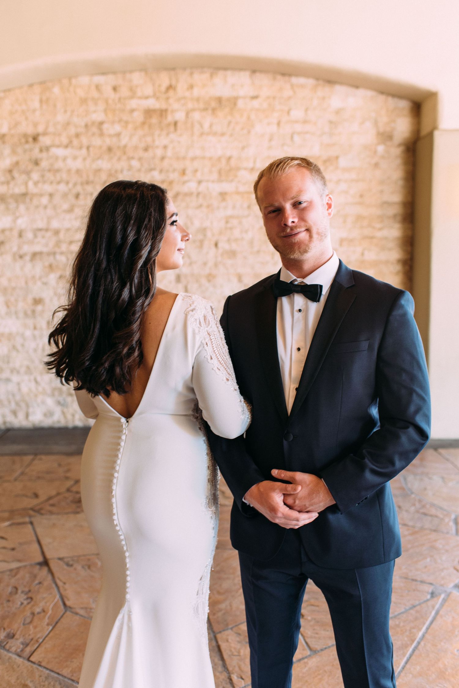 Bride in fitted white gown and groom in tuxedo pose against stone wall interior.