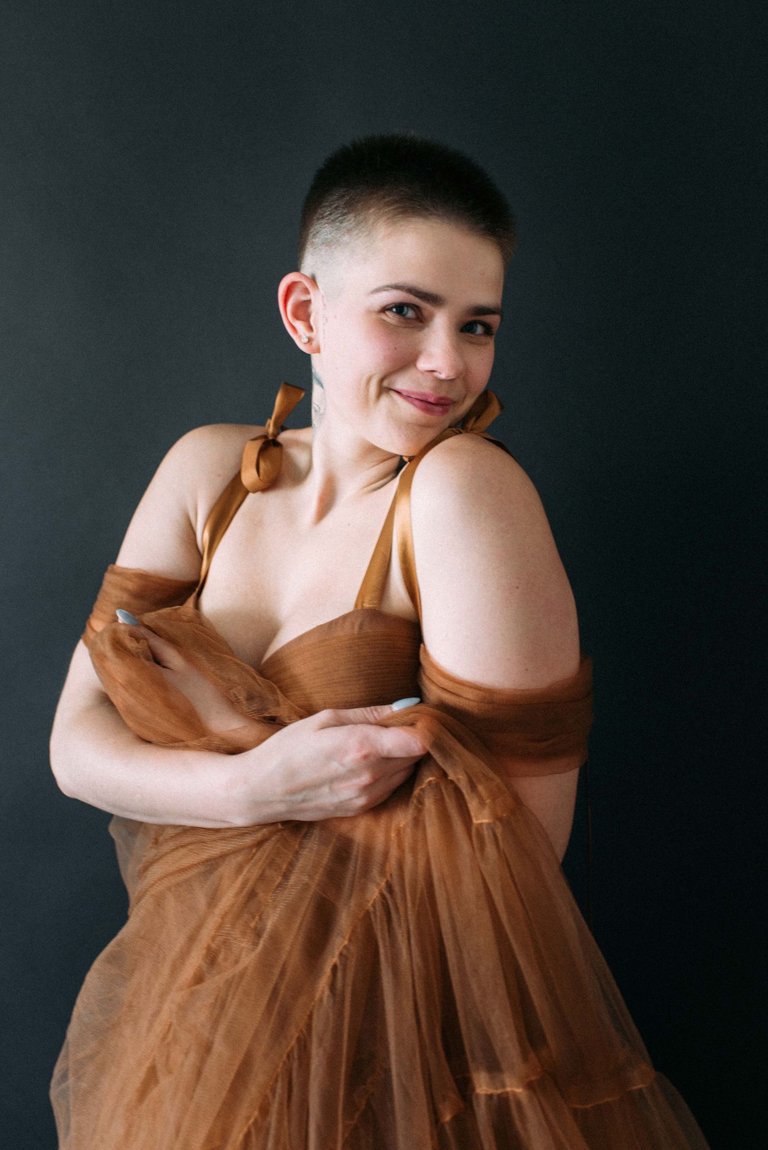 A figure in a flowing brown tulle dress poses against a dark background with a joyful expression.
