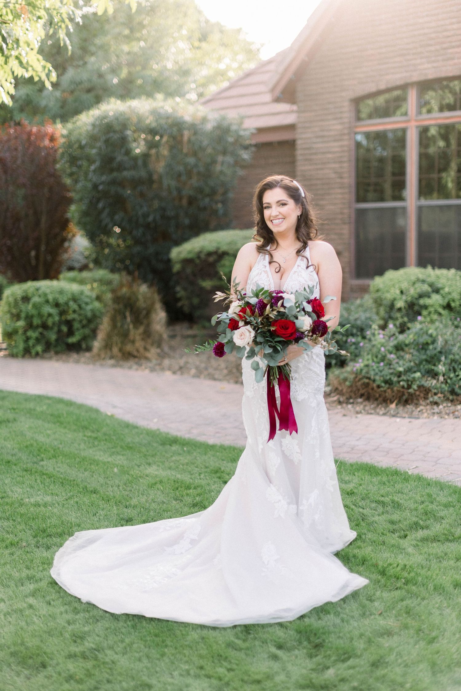 Full-length bridal portrait in lace gown holding red and white bouquet against brick building.
