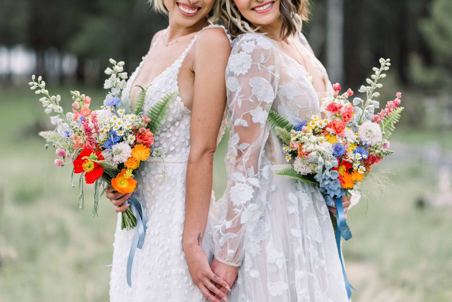 Two brides in white lace dresses hold vibrant wildflower bouquets while standing together in a forest setting.