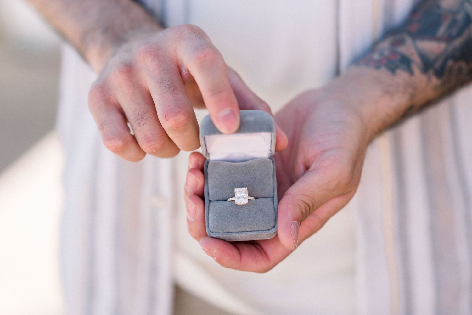 Hands holding a small gray ring box displaying an engagement ring against a white background.