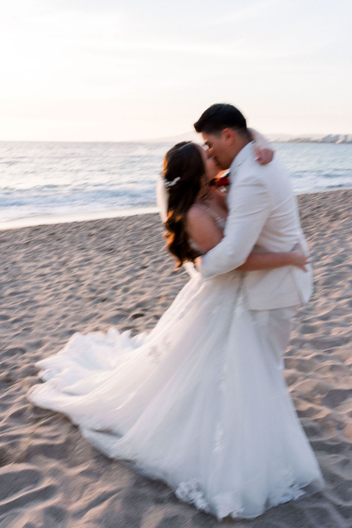 A romantic beachside wedding couple embraces during sunset with the bride's white dress flowing in the sand.