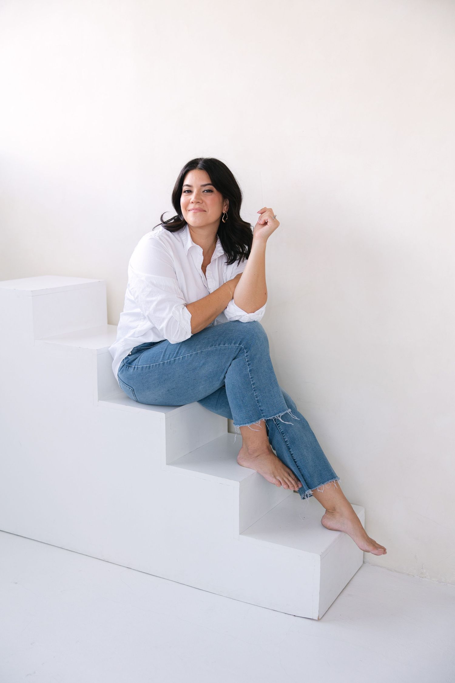 A figure in a white shirt and blue jeans poses artistically on a white geometric platform against a light background.