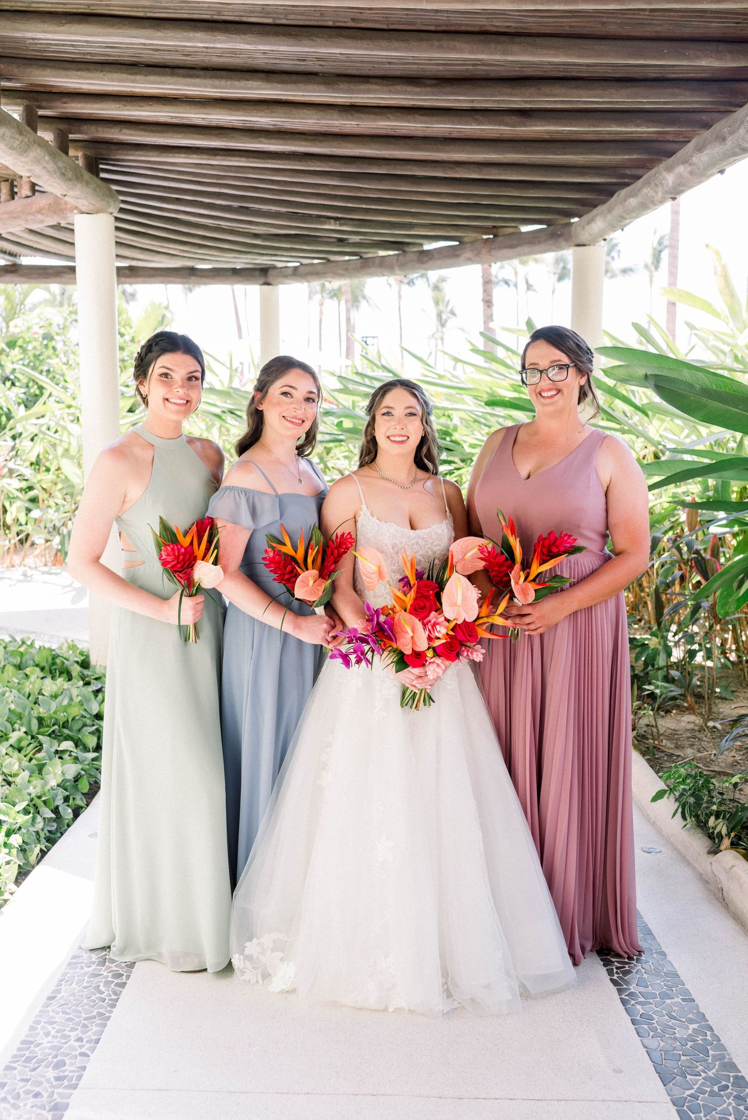 Bridal party posing under a covered walkway with tropical flowers and pastel colored dresses.
