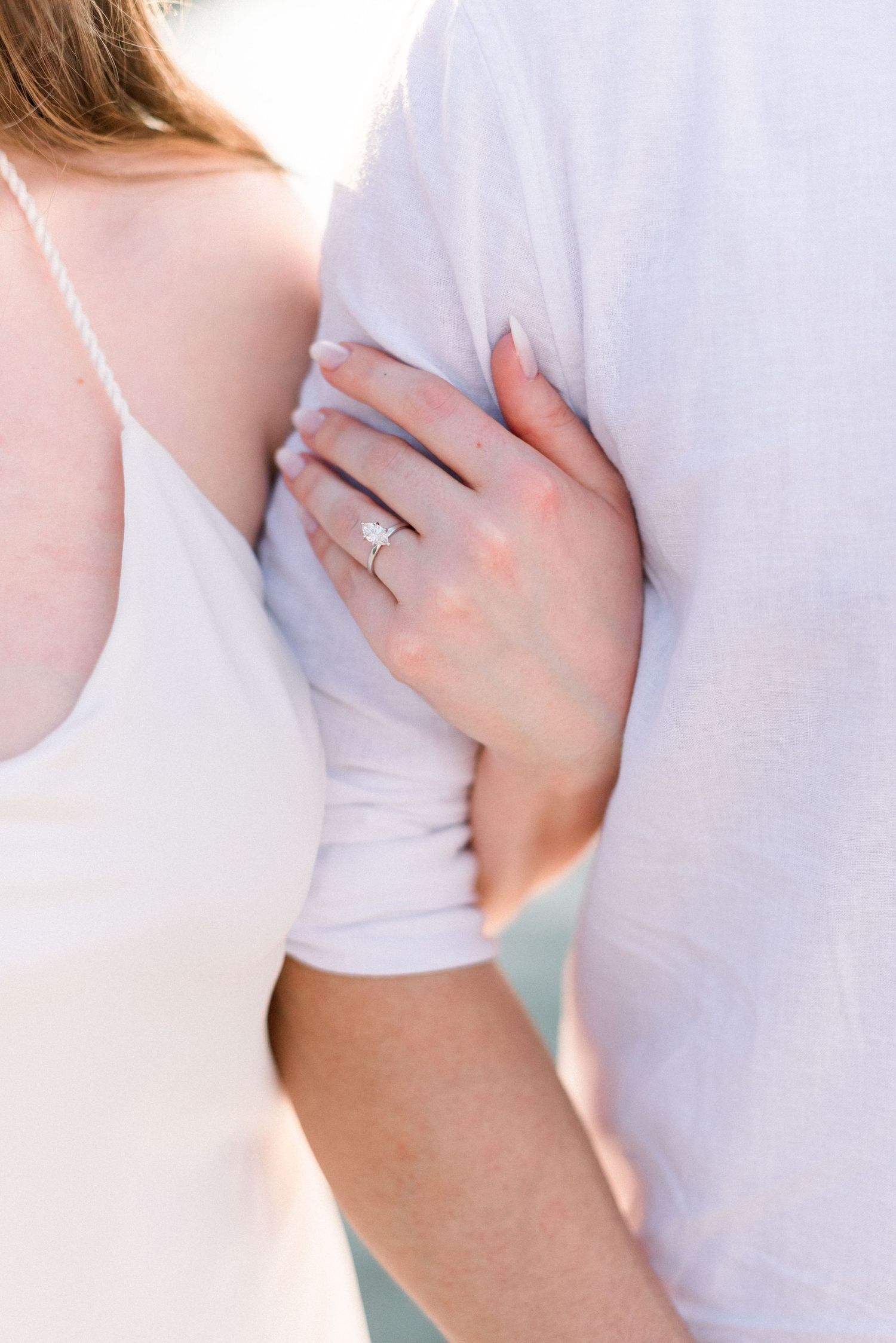 Close up of hands showing off an engagement ring against white clothing.