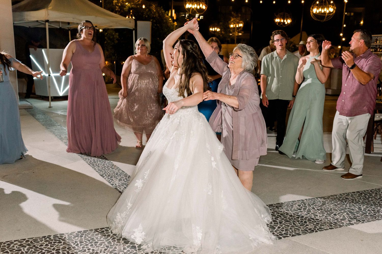 Wedding guests and the bride dancing together on a patterned floor at an outdoor evening celebration.