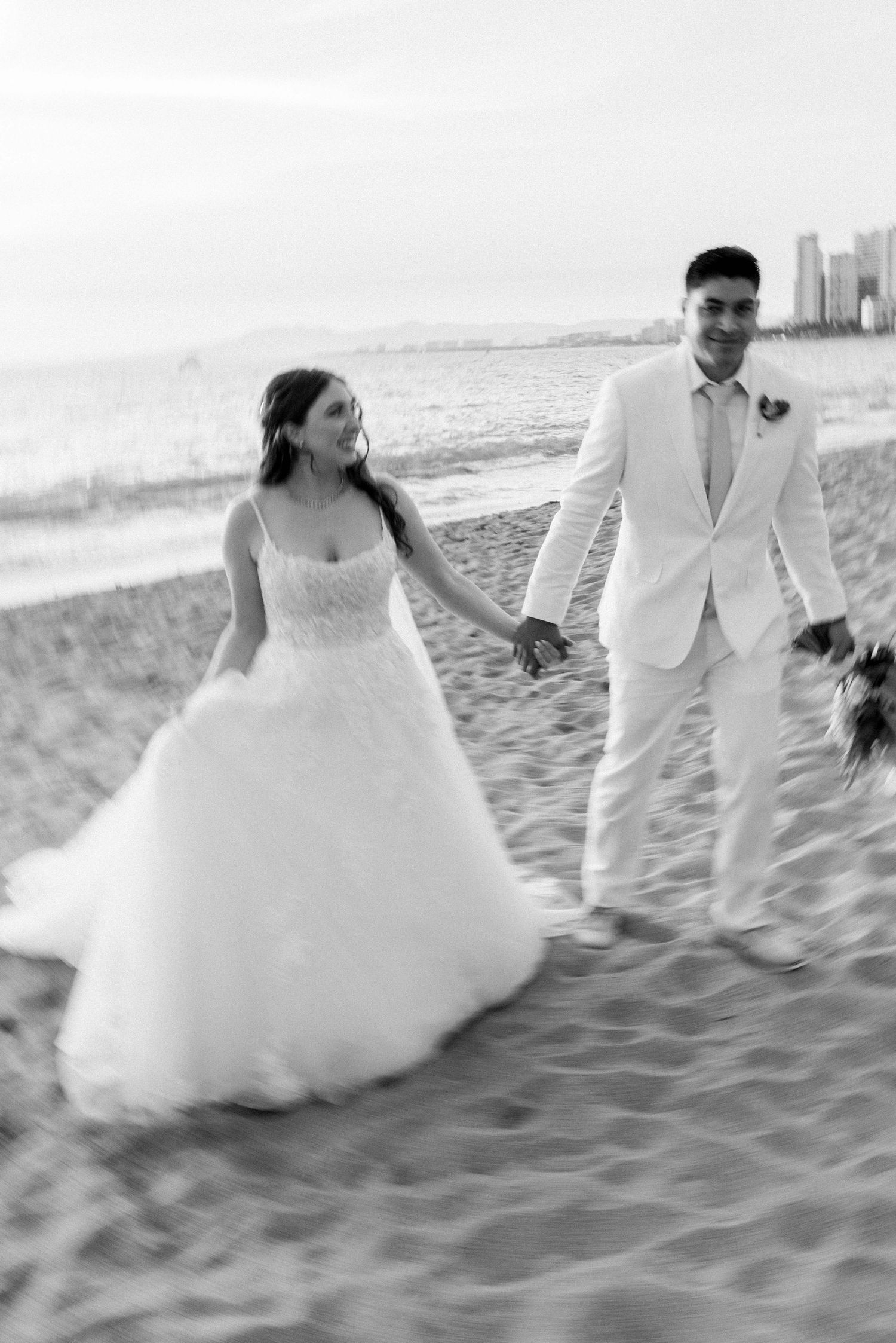 Joyful wedding couple walking on beach in black and white with cityscape in background.