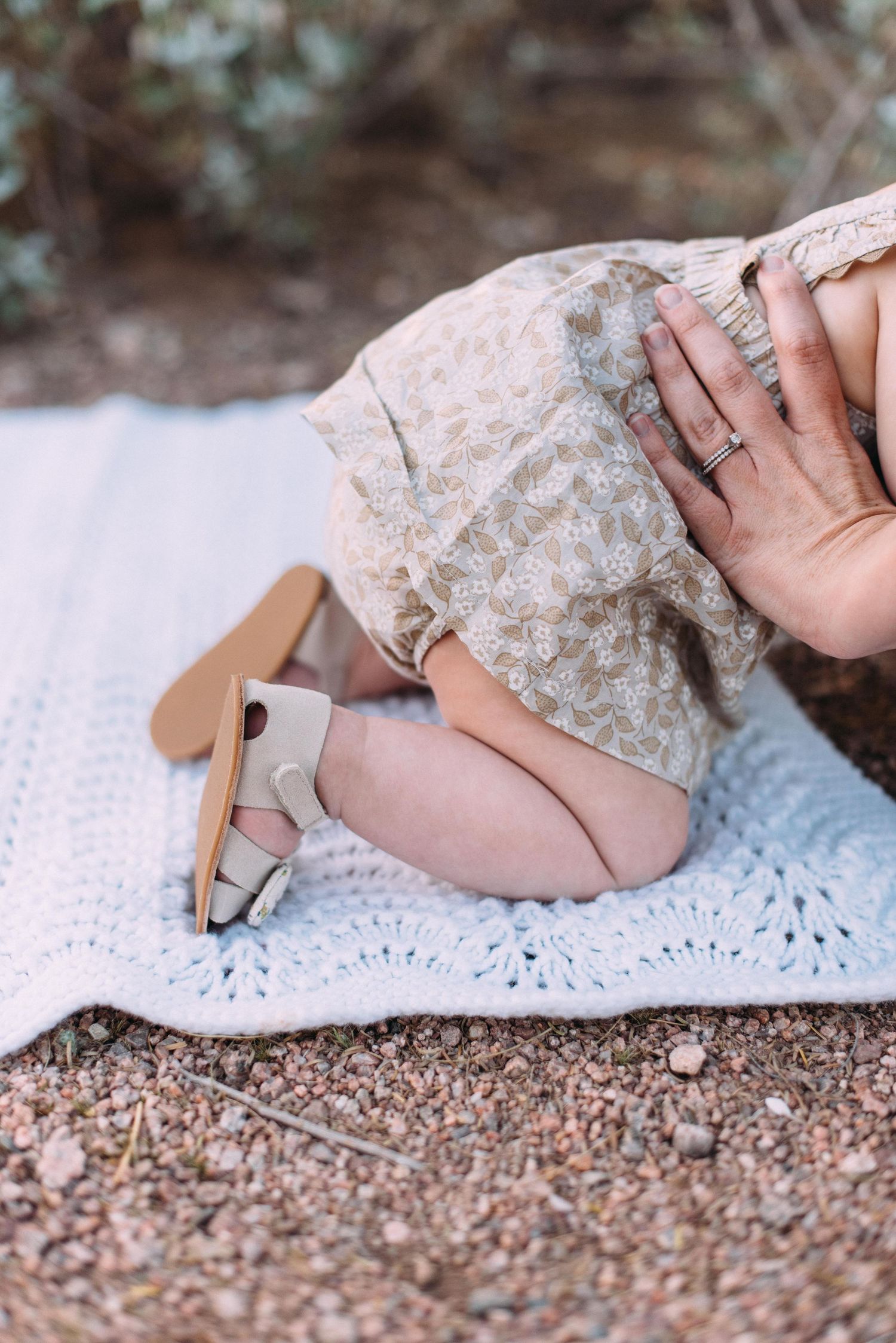A baby in a floral dress sits on a white lace blanket showing their hand wearing delicate jewelry.