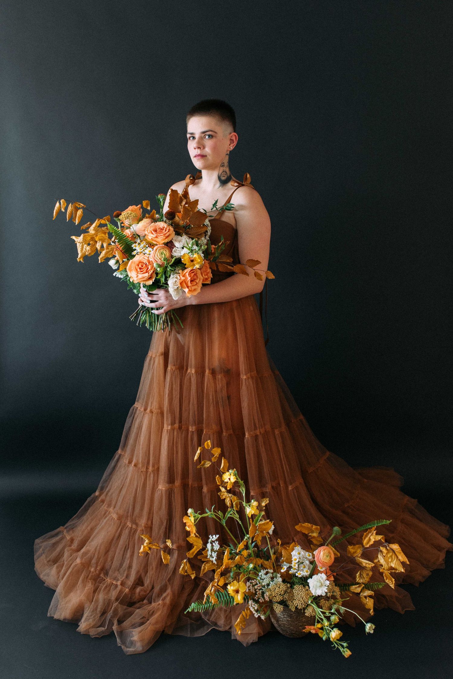 Portrait in flowing brown tulle gown holding autumn-toned floral arrangement against dark backdrop.
