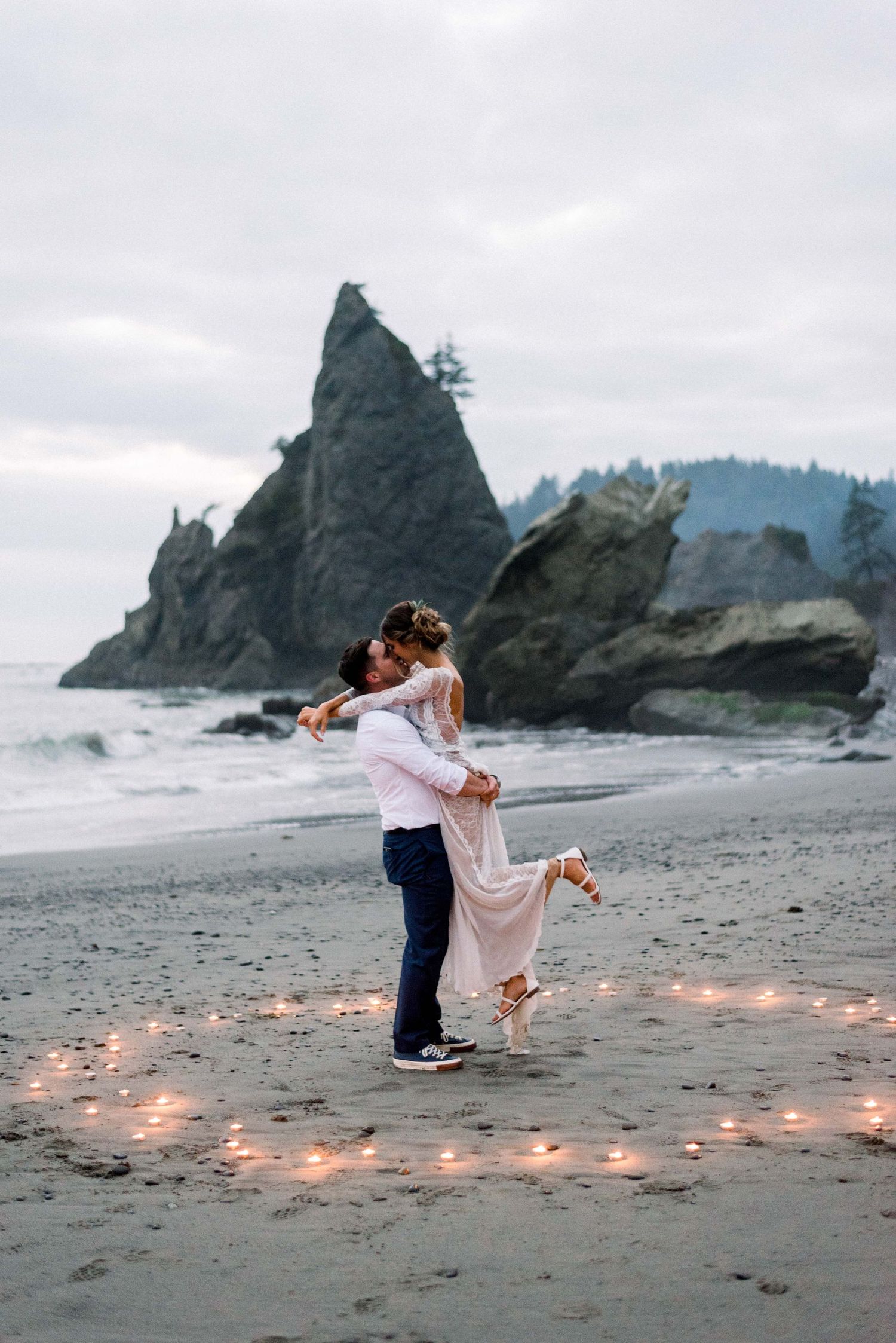 Couple dancing on beach with candlelit path and dramatic coastal cliffs in background.