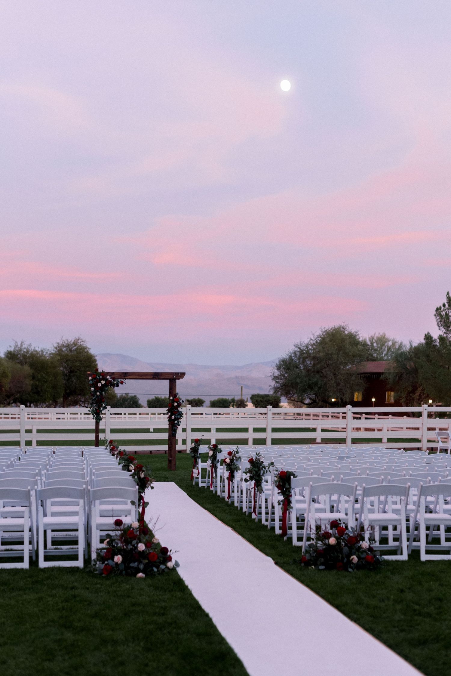 Outdoor wedding ceremony setup with white chairs and floral arch under pink sunset sky.