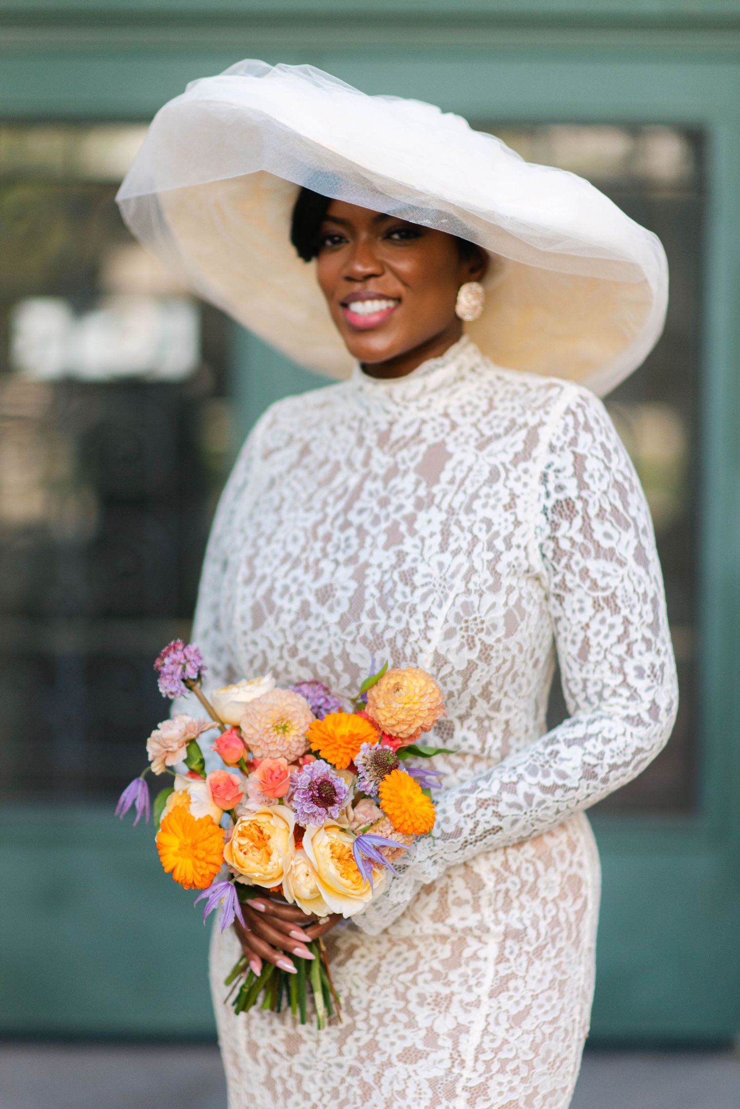Elegant bride in white lace dress and dramatic wide-brimmed hat holds vibrant orange and purple floral bouquet.