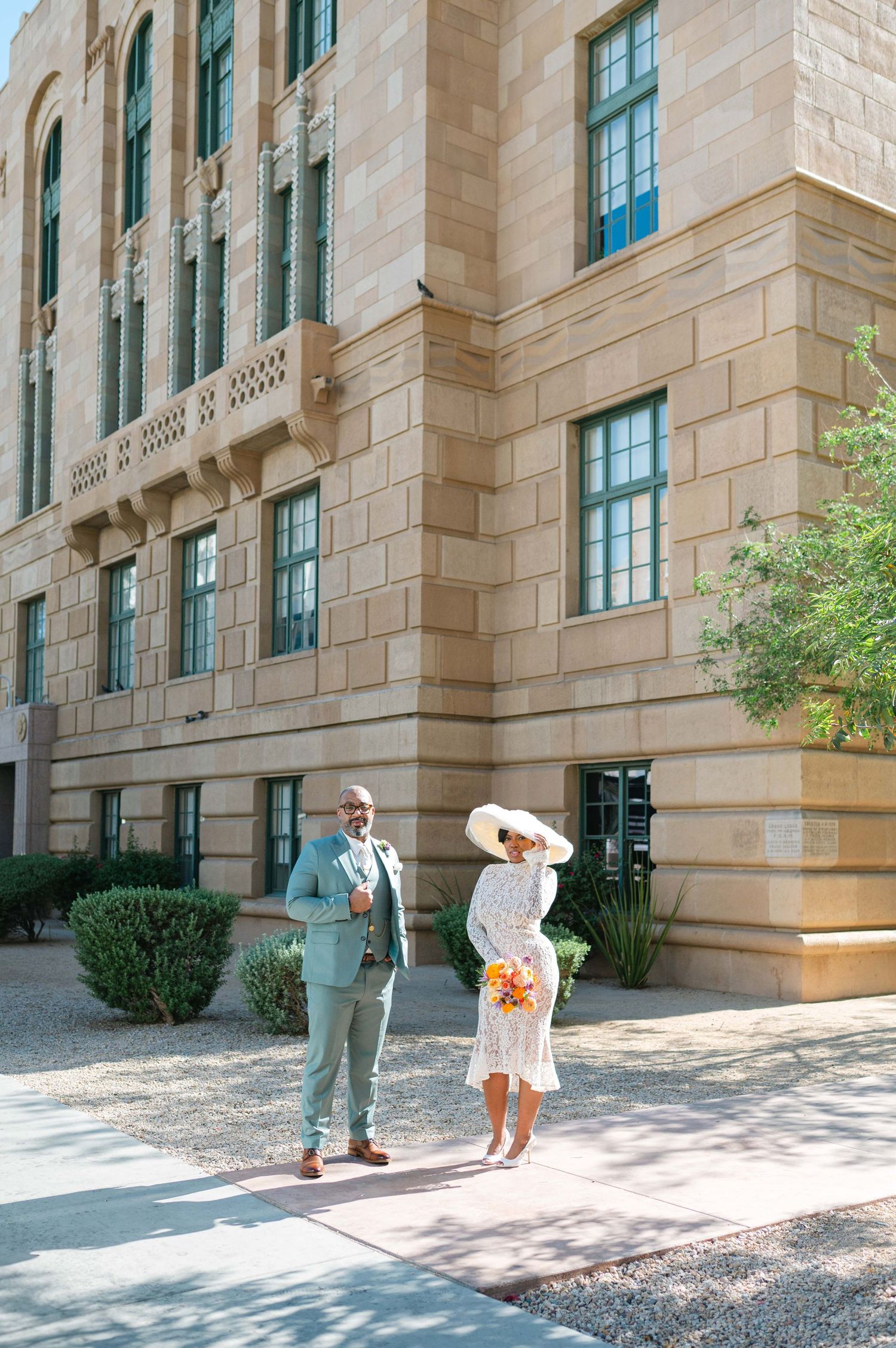 A couple poses in sequence outside a historic sandstone courthouse building with flowers.