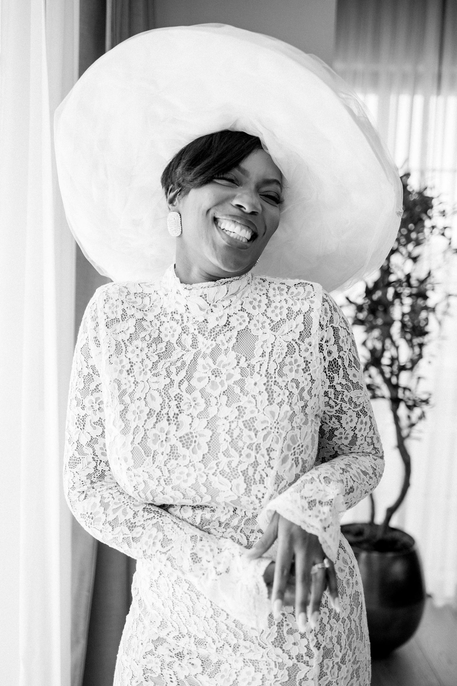 Bride wearing a stunning lace dress and oversized white hat smiles joyfully in black and white portrait.