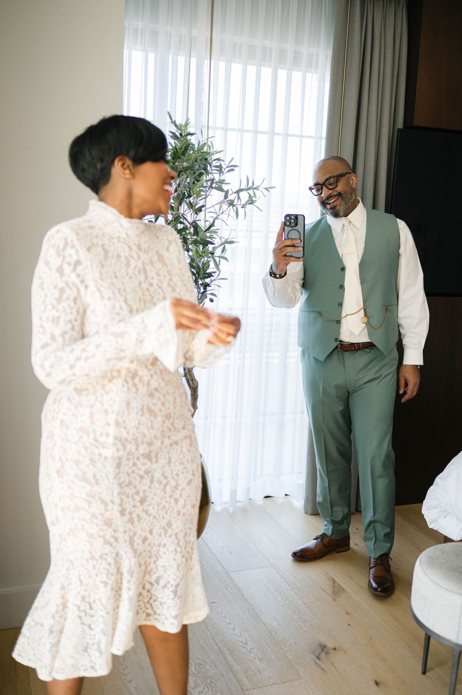 A couple in a bright room, with one wearing a white lace dress and another in a sage green suit taking a photo.