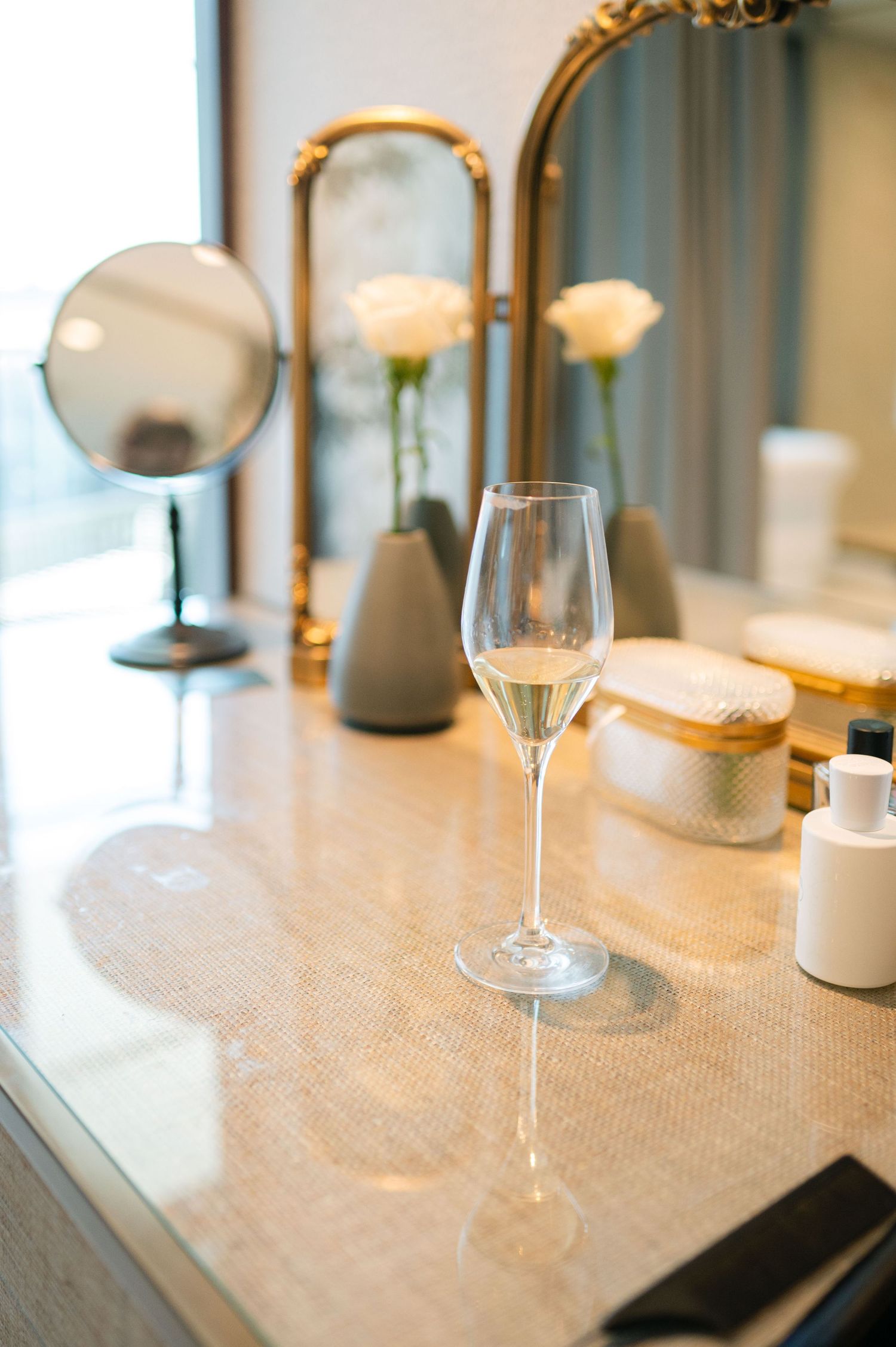 A glass of champagne sits on a vanity table with roses in vases and a circular mirror in an elegant setting.