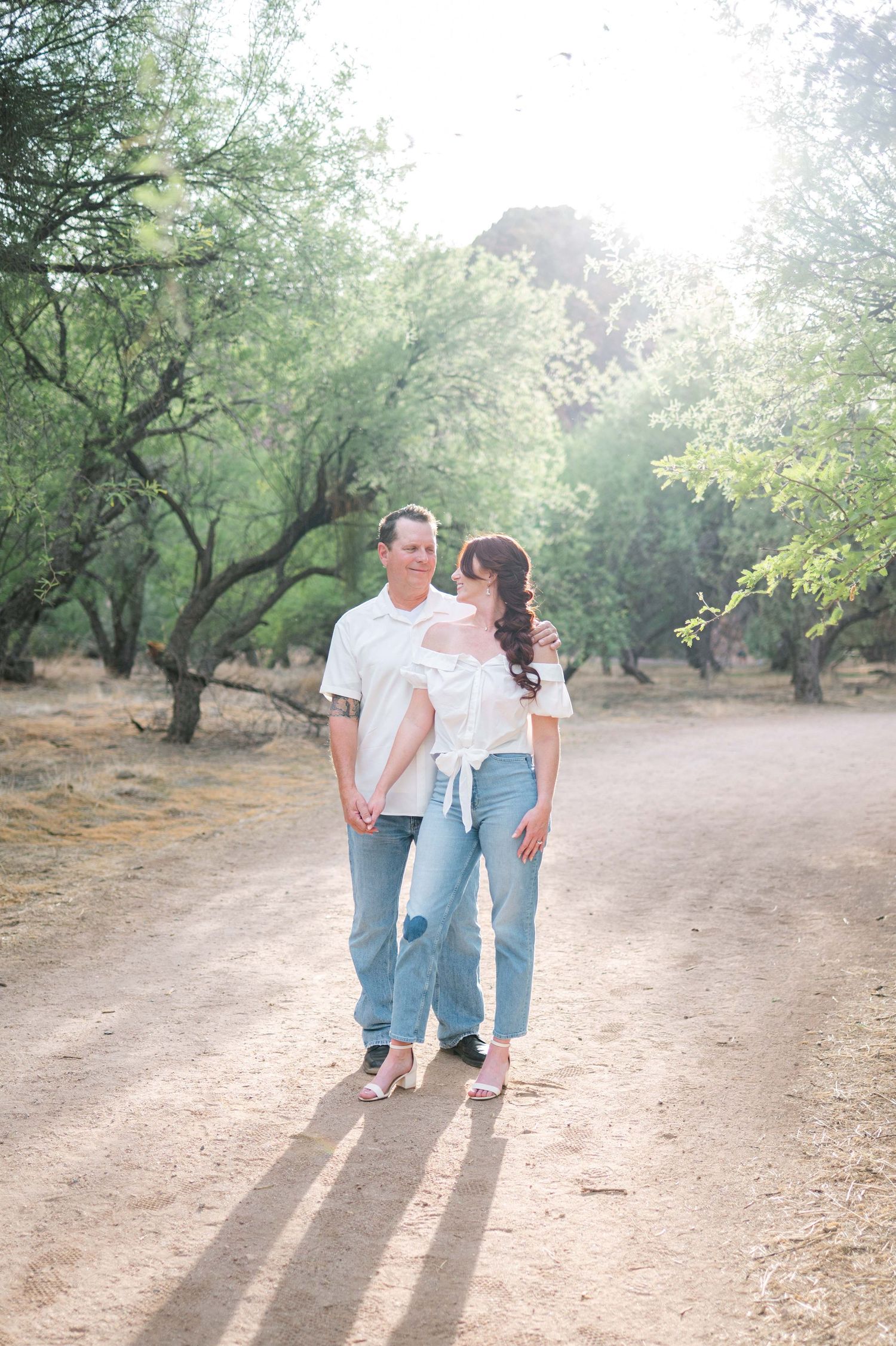 A couple walks hand in hand down a sunlit dirt path through an orchard during a romantic outdoor photoshoot.