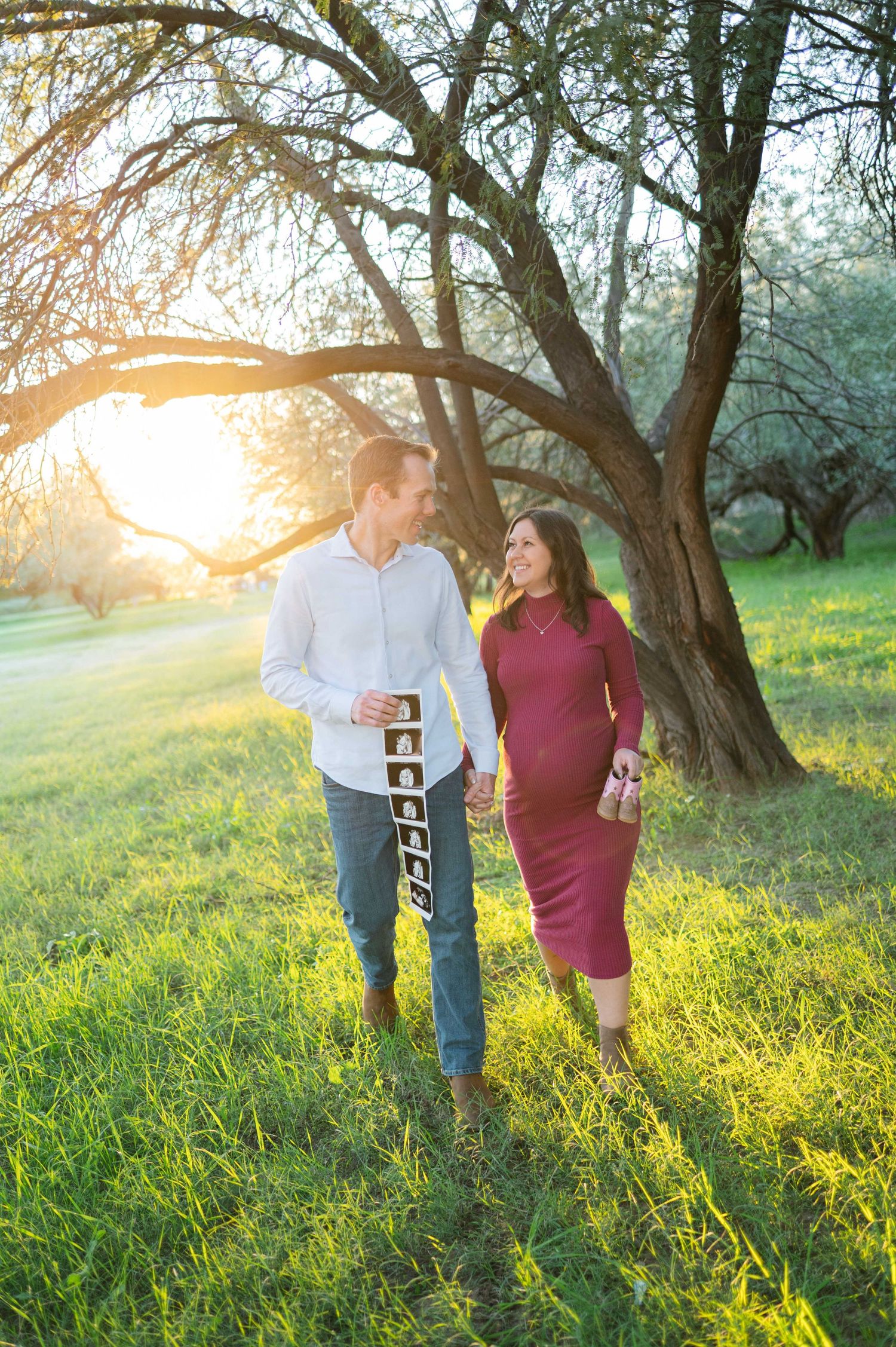 A couple walks hand in hand through a sunlit meadow with blooming trees during golden hour.