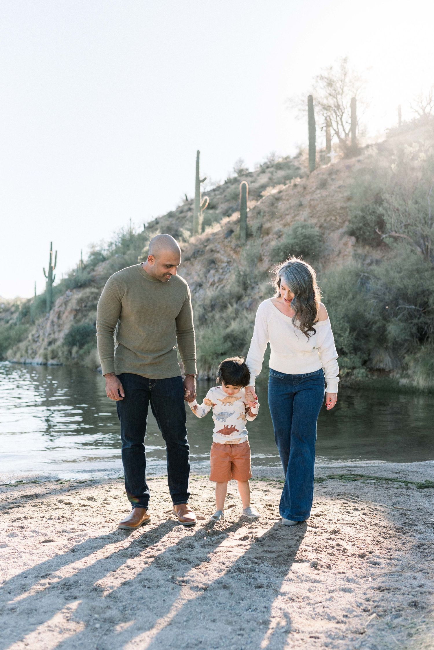 Family walking together along a desert lake shore with saguaro cacti in the background creating long shadows at sunset.