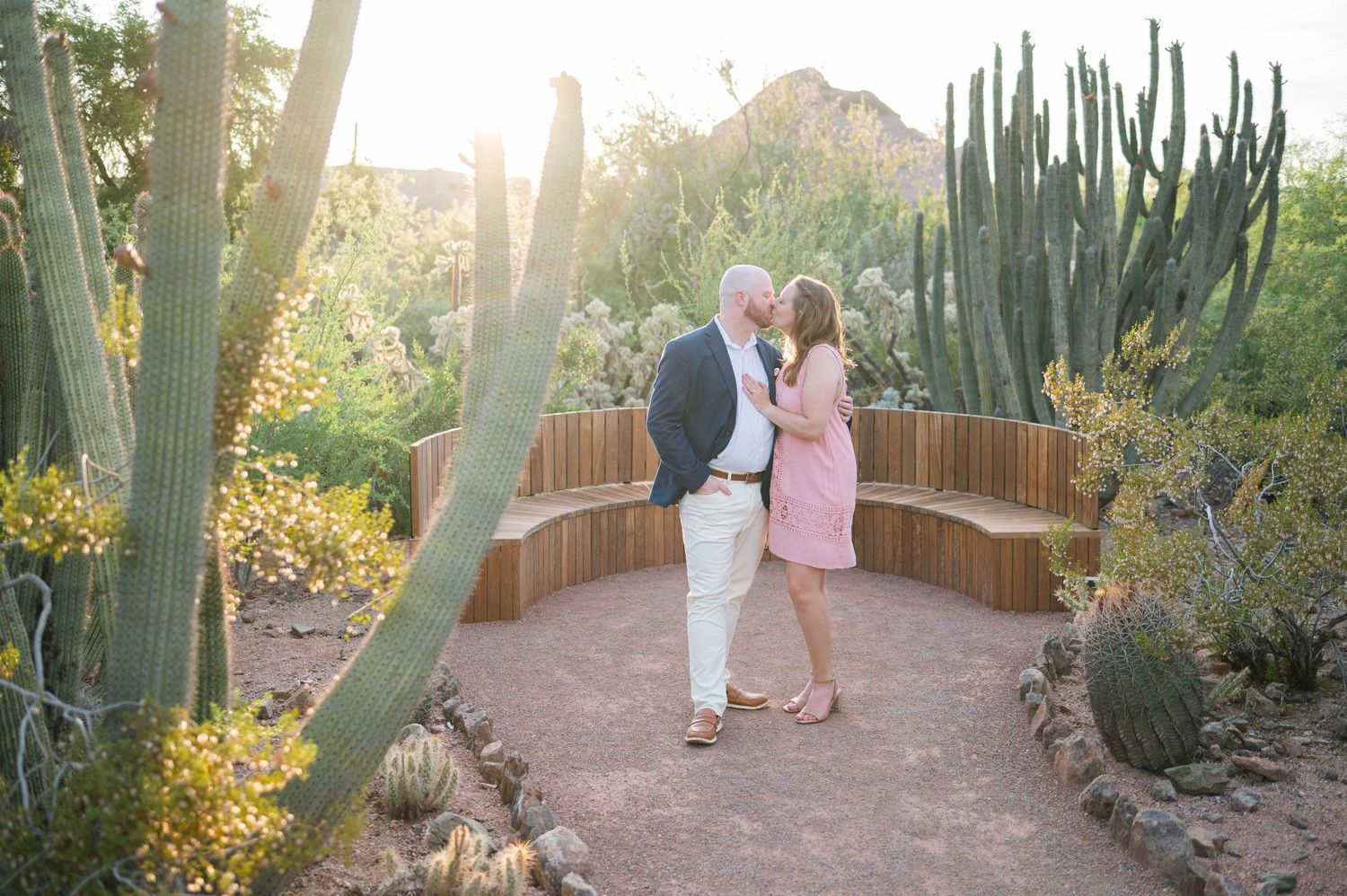 A couple shares a romantic moment on a curved pathway surrounded by desert cacti and succulents at sunset.