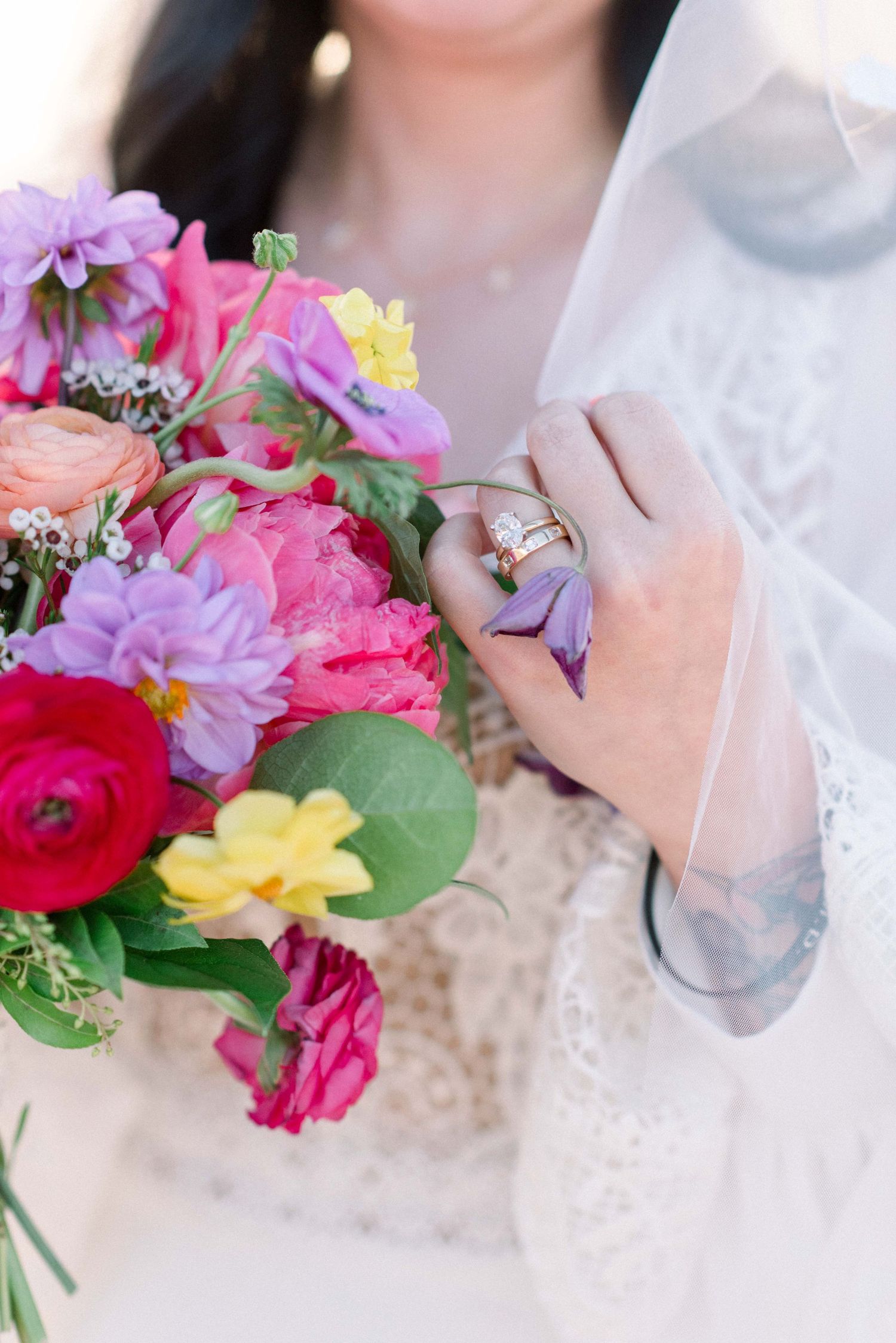 Colorful wedding bouquet with pink, purple, and yellow flowers held by hands in white lace.