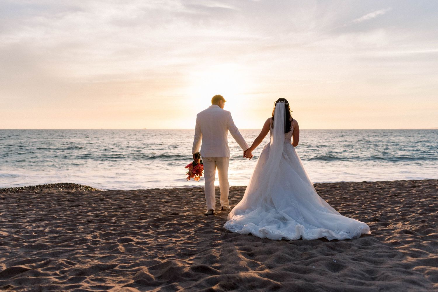 Wedding couple silhouetted against golden sunset holding hands on beach with flowing white gown.