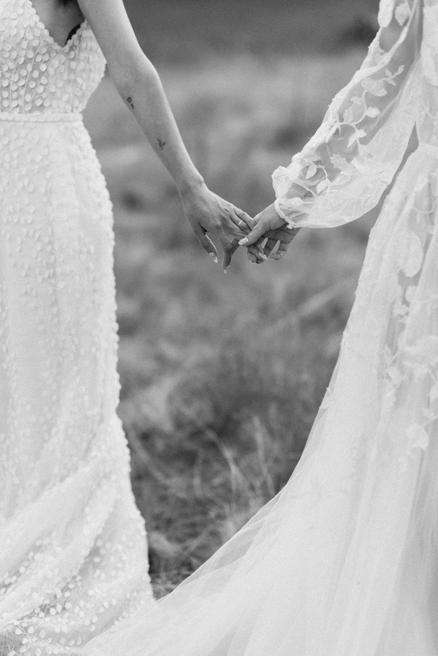 Black and white detail of two brides holding hands, showing the flowing fabric of their wedding gowns.