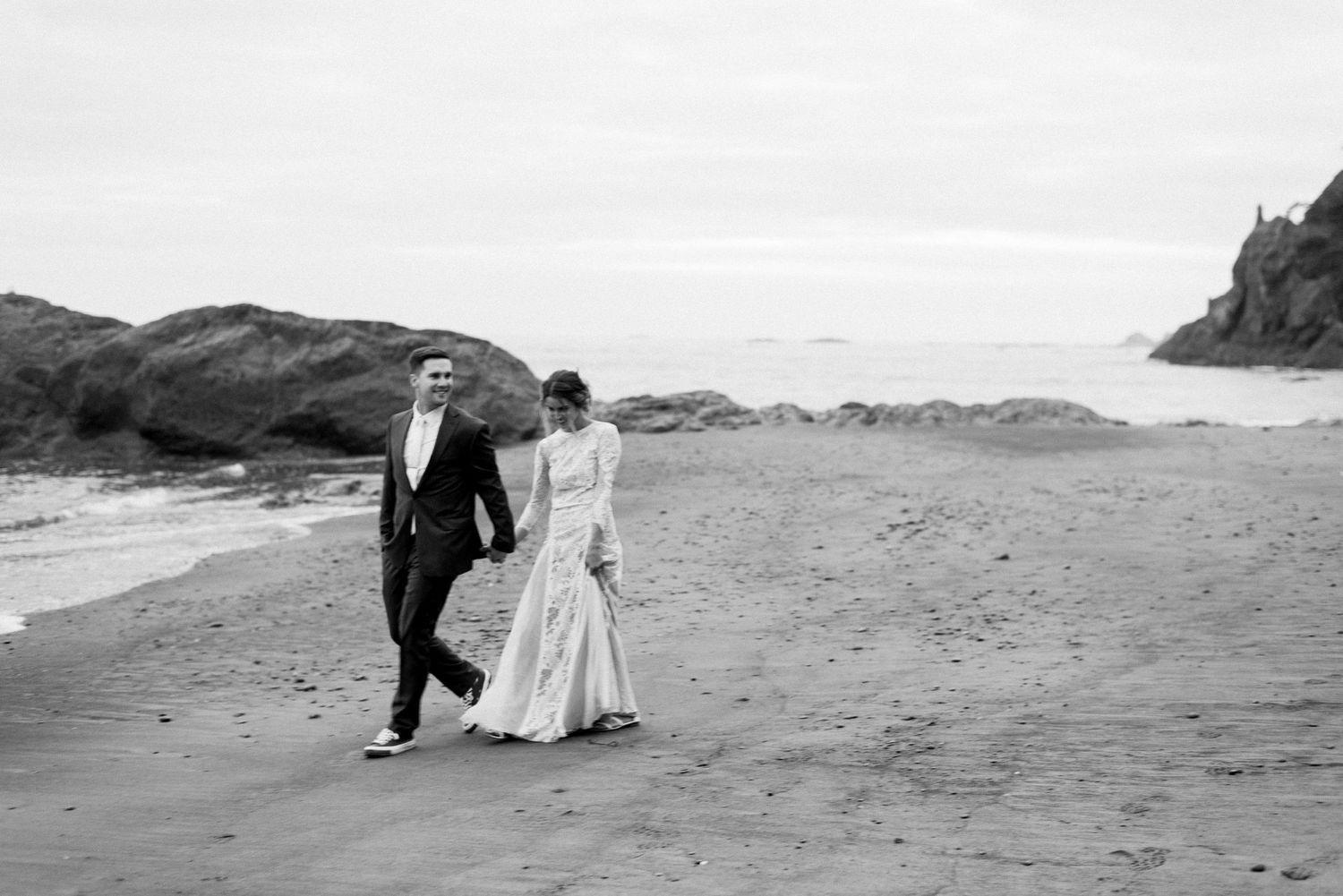 Black and white photo of newlyweds walking along a misty beach with coastal rocks.