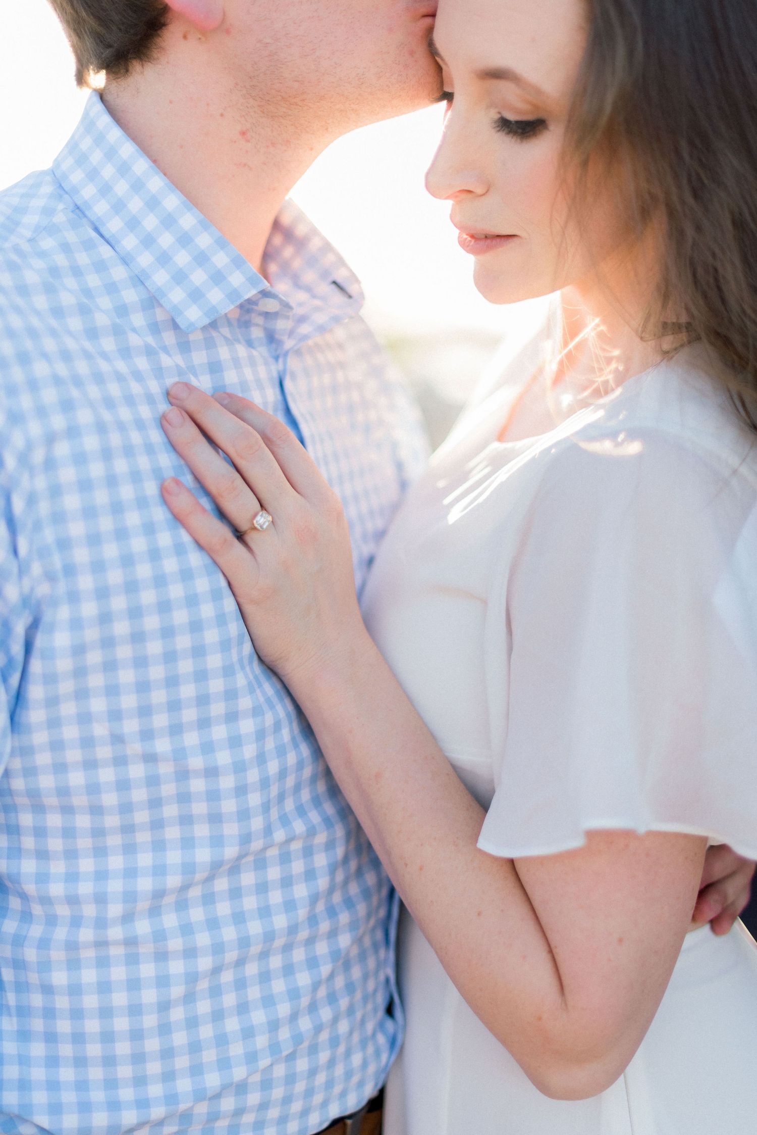 A romantic close-up photo showing a white gingham shirt and delicate embrace in soft lighting.