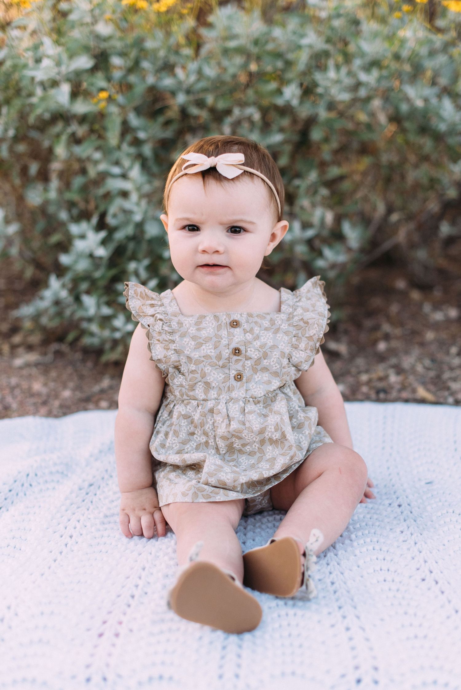 Baby in floral dress and bow sits on white blanket outdoors.