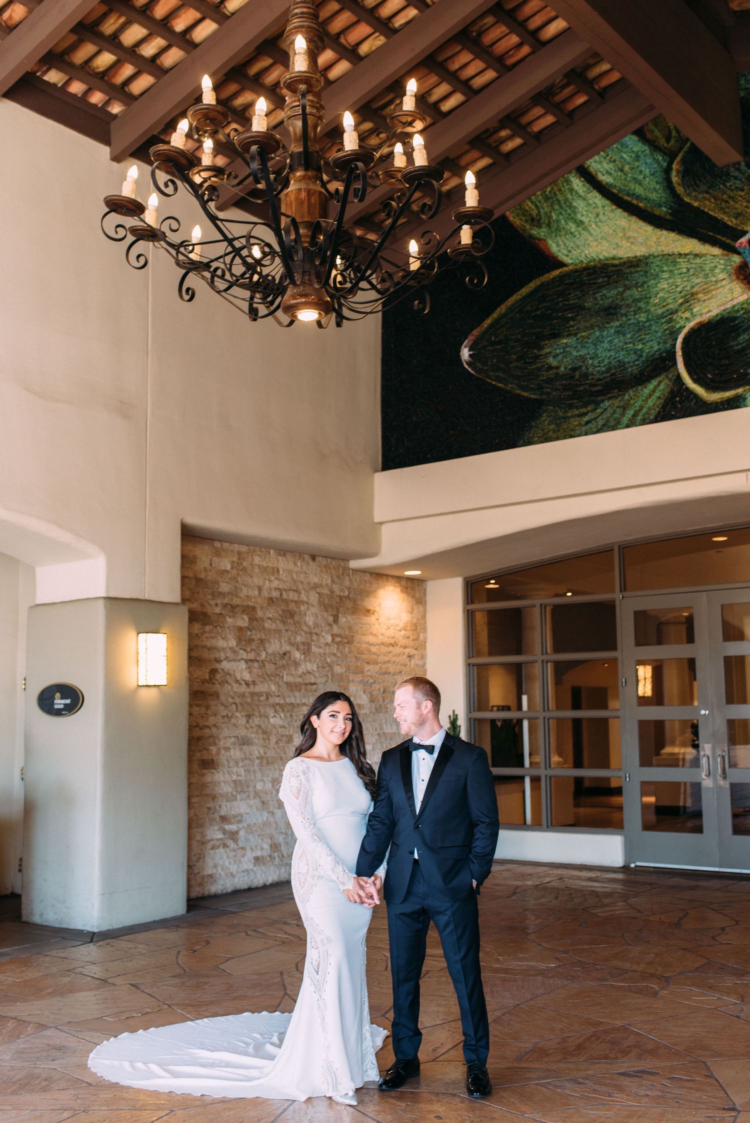 Elegant couple poses beneath ornate chandelier in grand entrance hall.