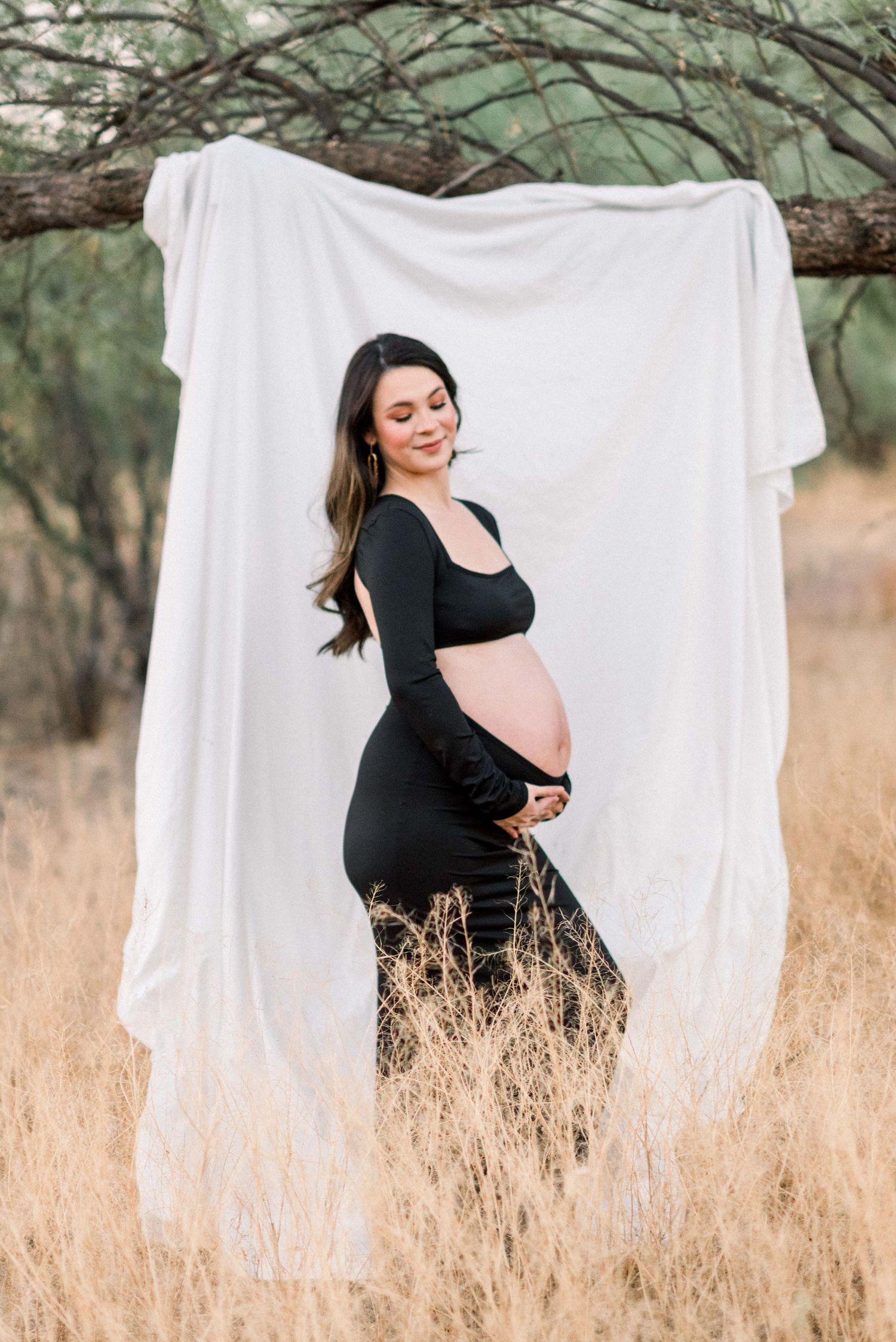 Joyful maternity portrait against white backdrop with woman in black dress surrounded by desert scenery.