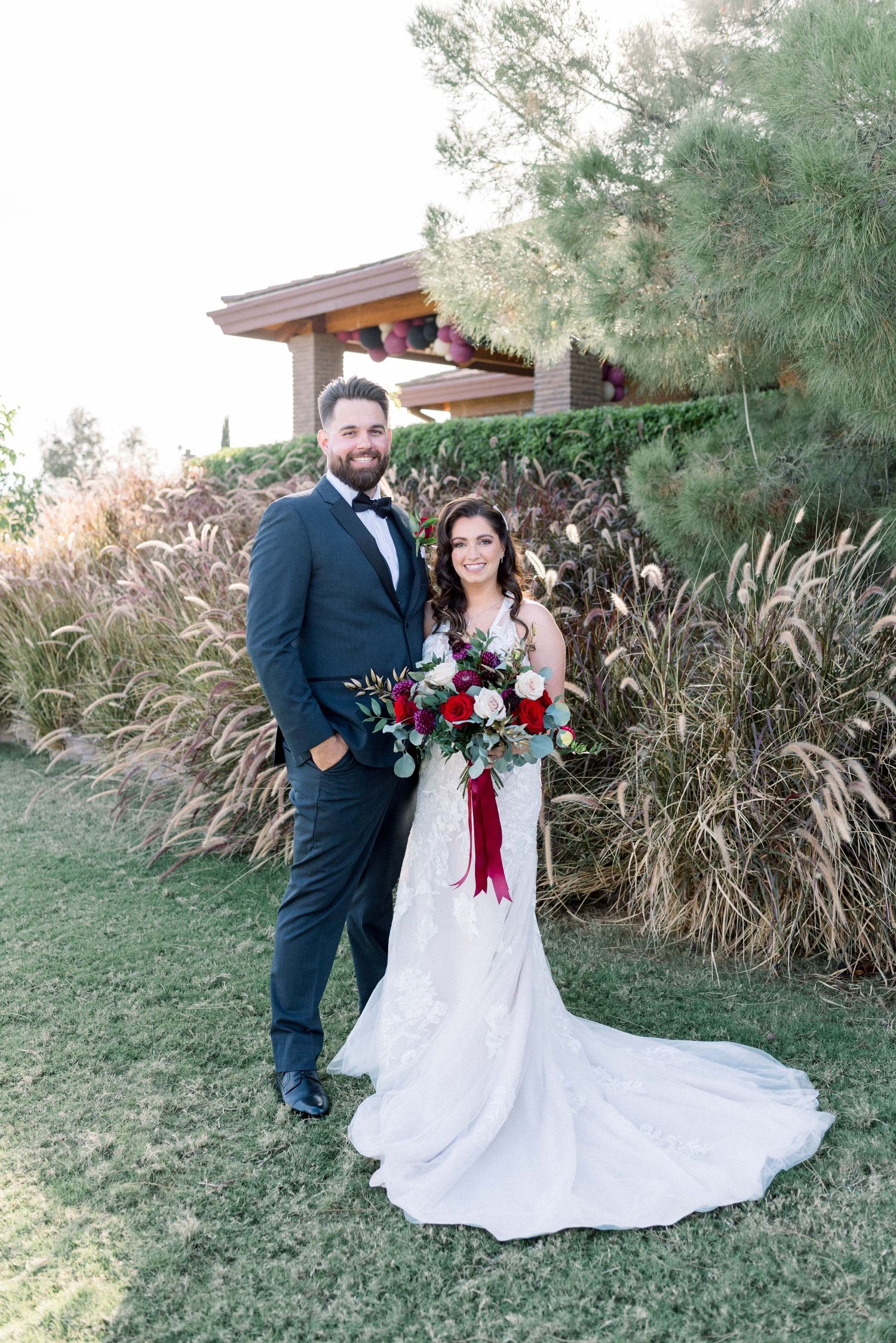 Newlyweds pose outdoors with red bouquet against ornate architecture and natural landscaping.
