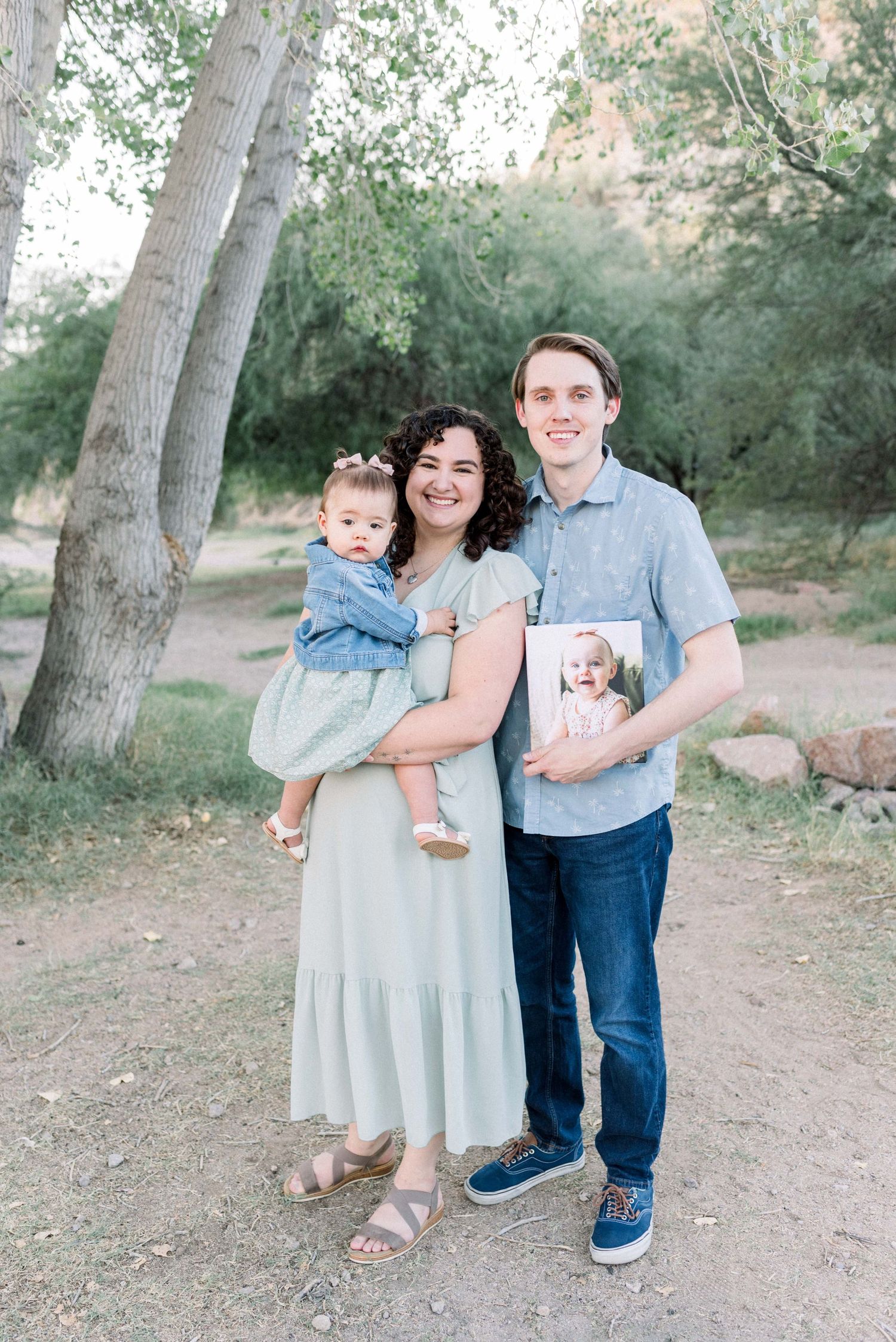 A family of three poses together outdoors in coordinating blue and neutral outfits against a backdrop of trees.
