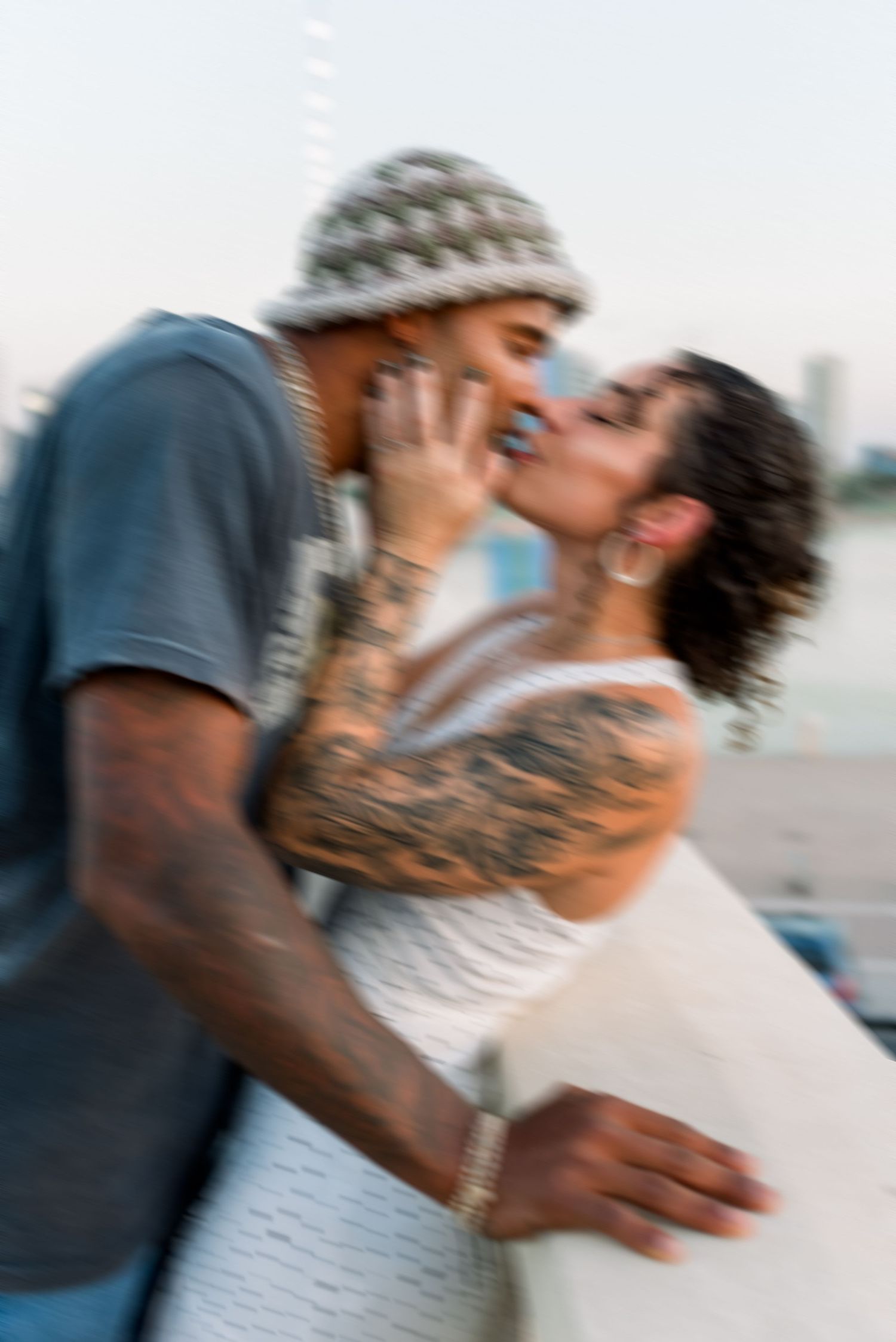 A romantic couple sharing a tender kiss outdoors with visible tattoos and a beanie hat in a candid moment.