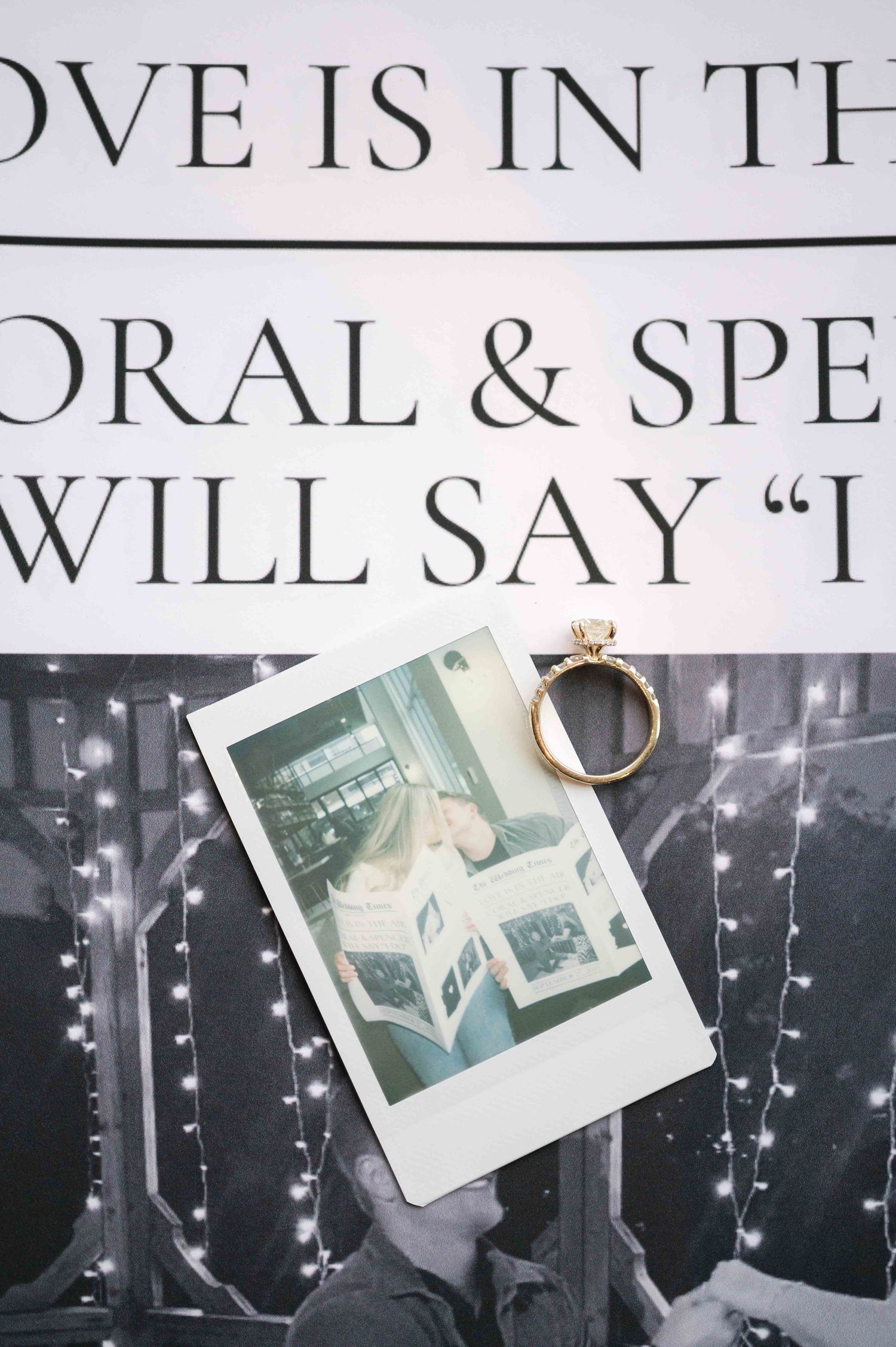 A romantic engagement ring rests on a polaroid photo against a backdrop with text and twinkling lights.