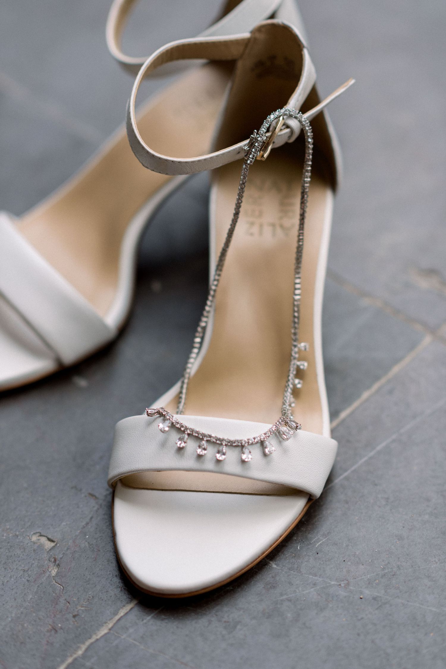 Elegant white wedding sandal with crystal embellishments and ankle strap on stone surface.