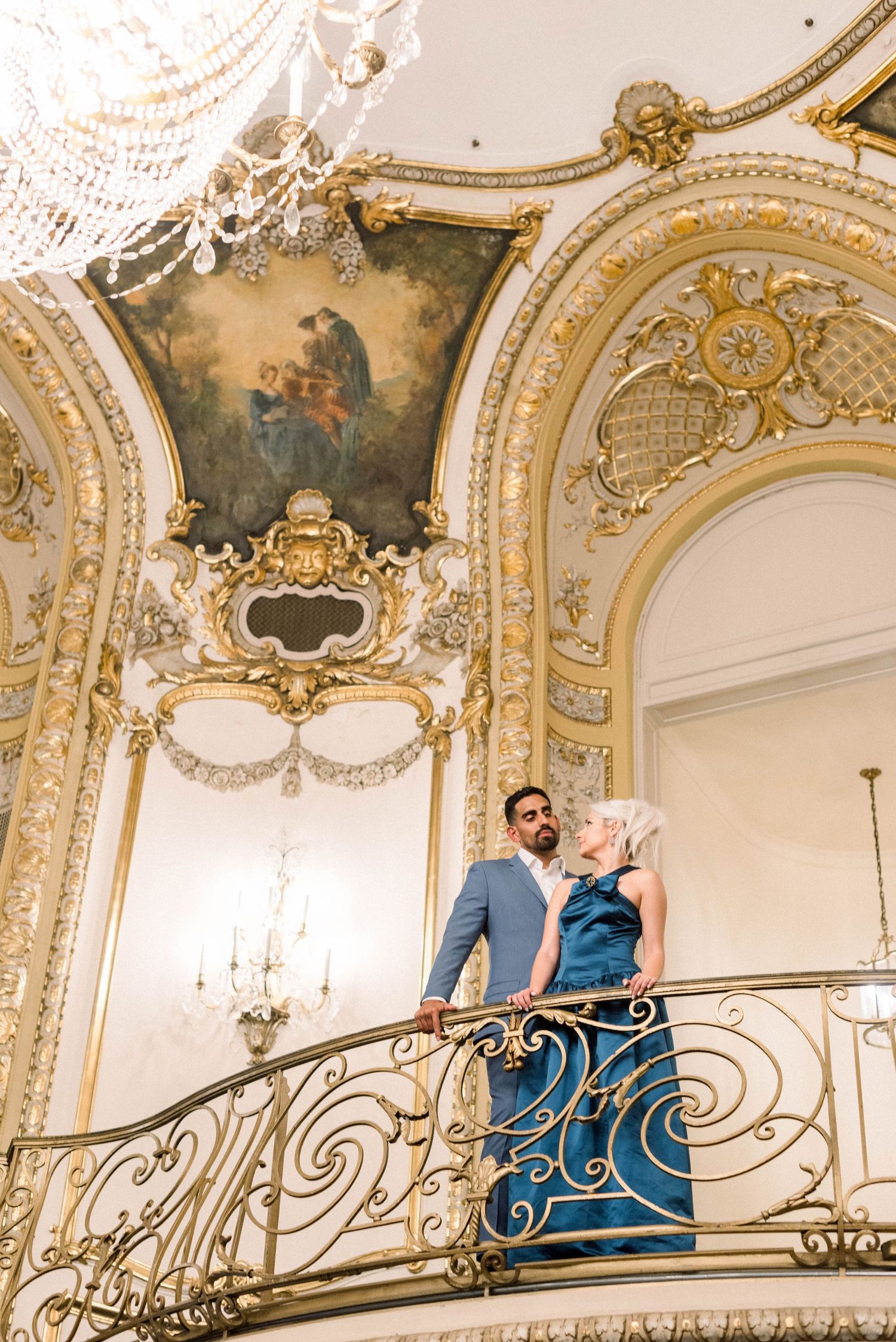 Couple poses on ornate balcony with baroque ceiling details and chandelier.