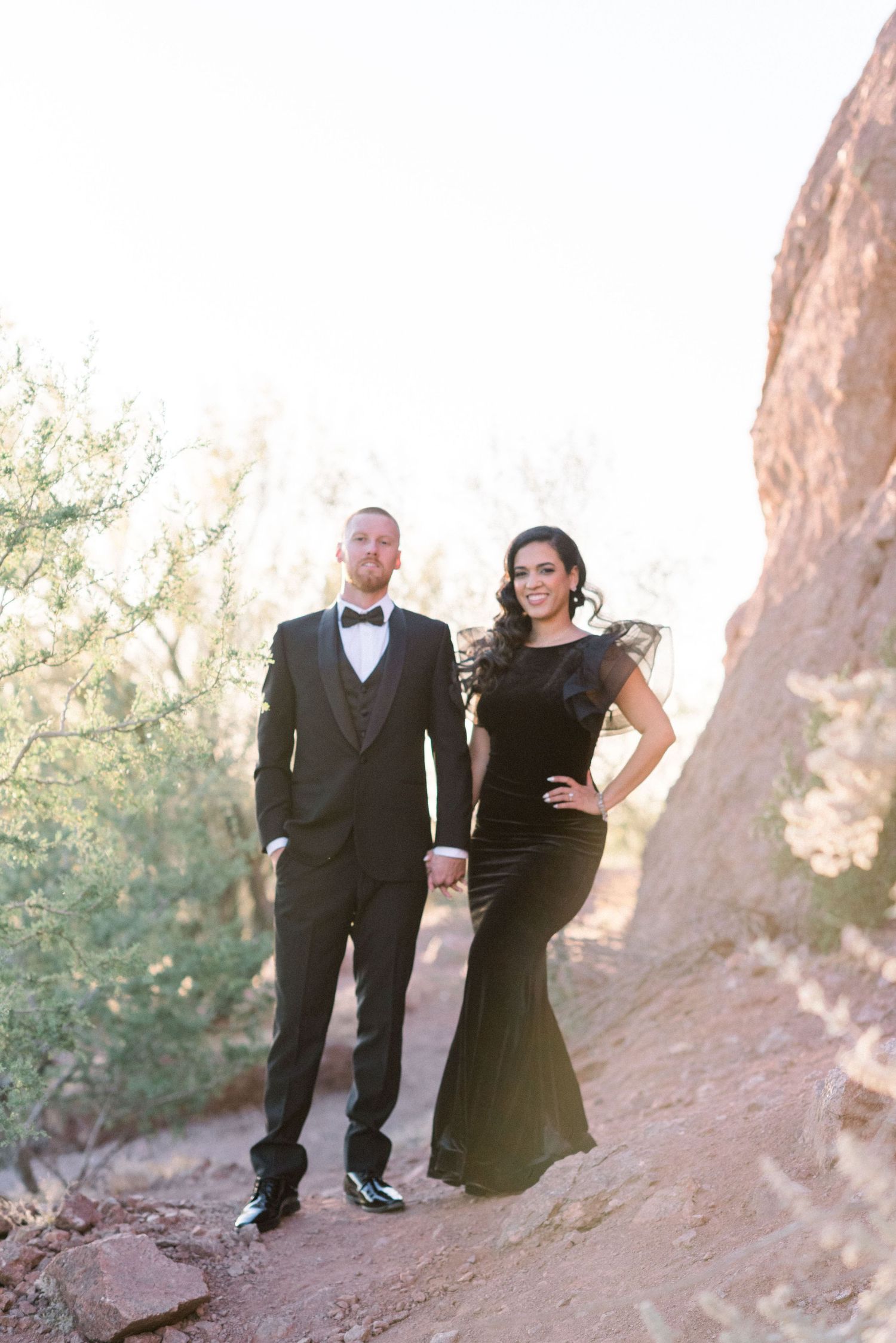 Formal couple posing in black tie attire against desert mountain backdrop.