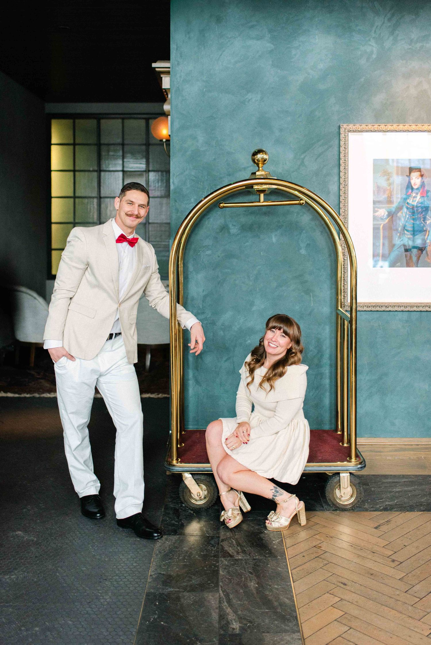 A couple poses playfully with a vintage hotel luggage cart against a teal wall.