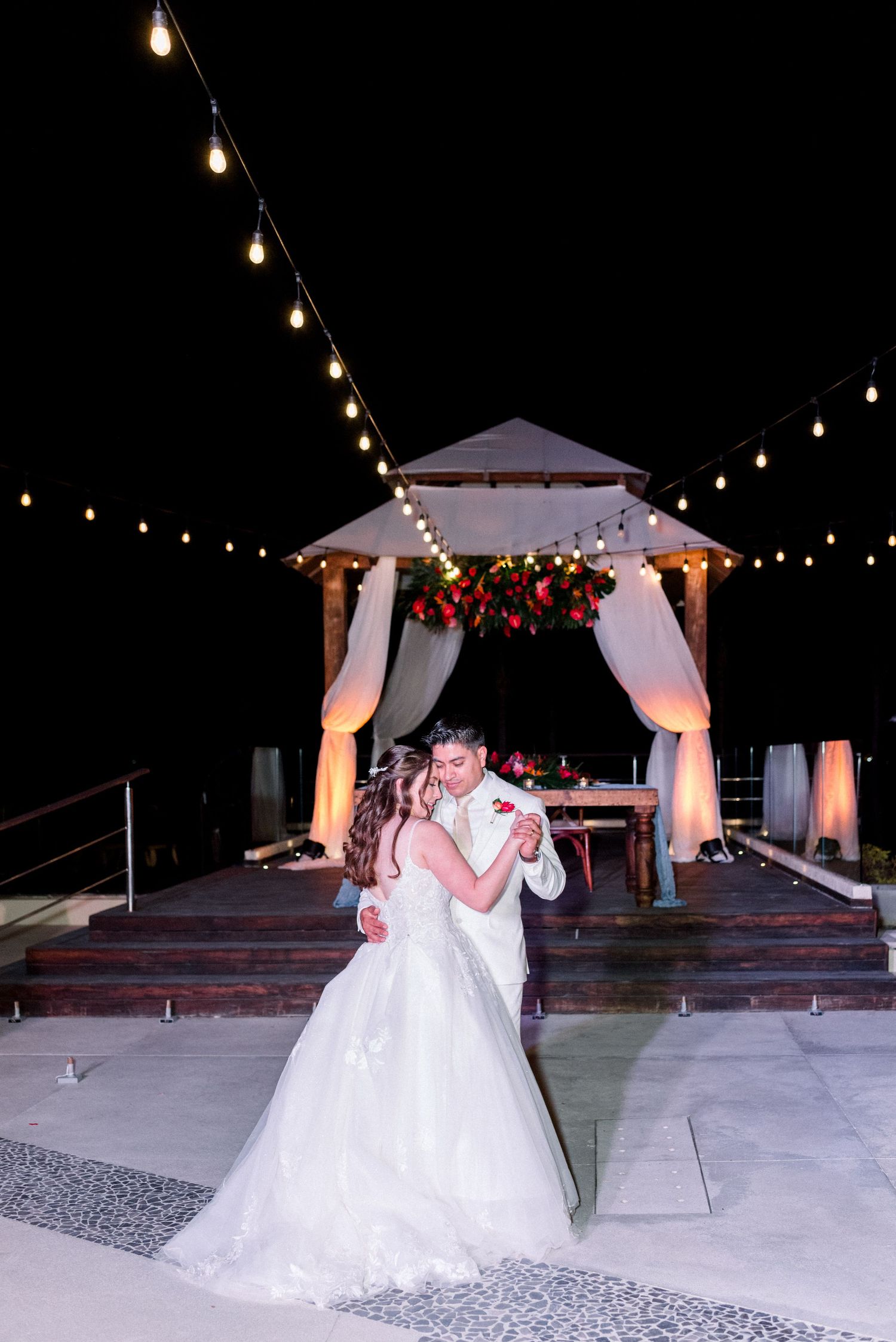 Couple dancing under illuminated gazebo decorated with fabric and string lights.