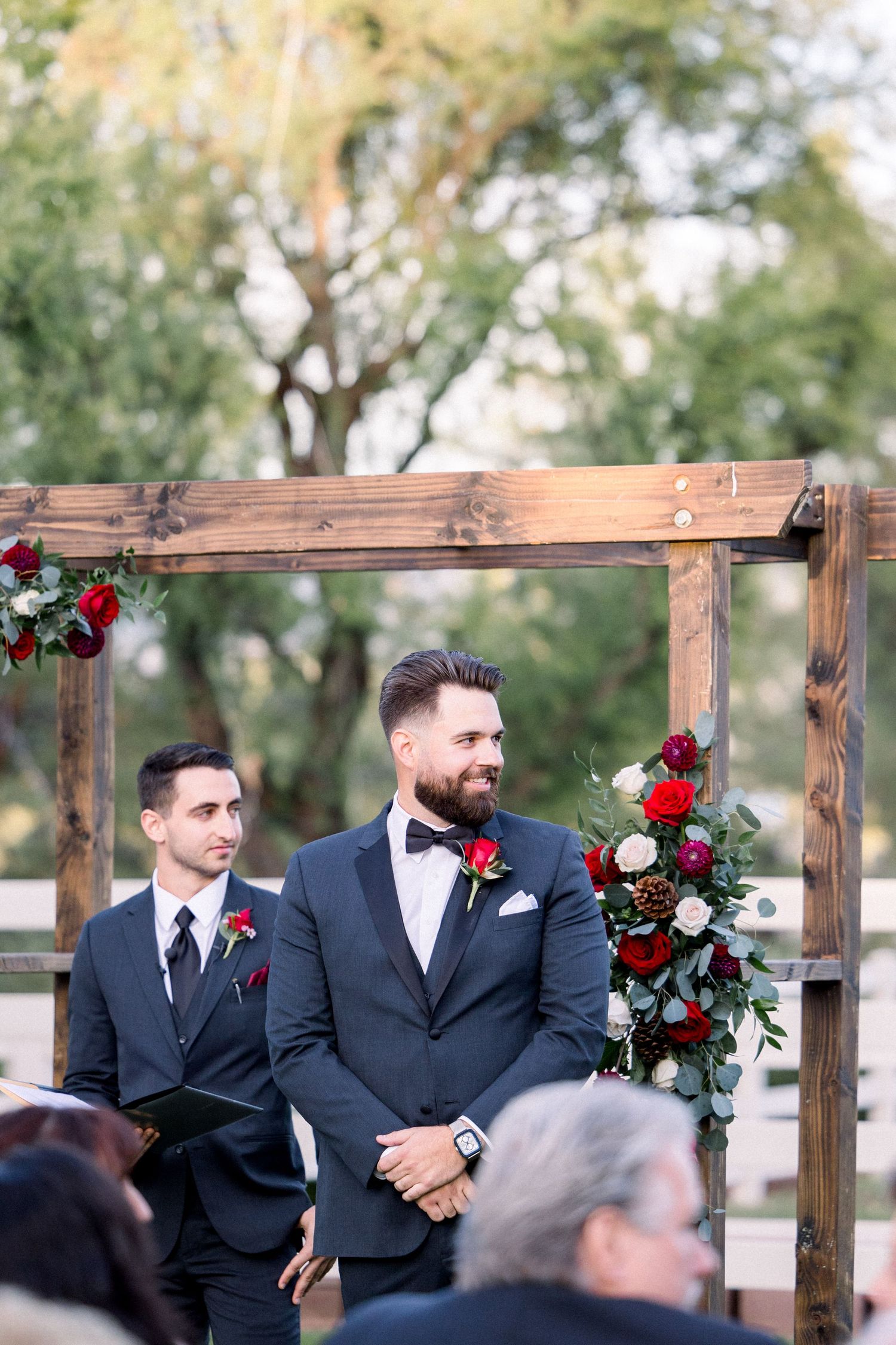 A rustic wooden wedding arch decorated with red roses and greenery stands at an outdoor ceremony.
