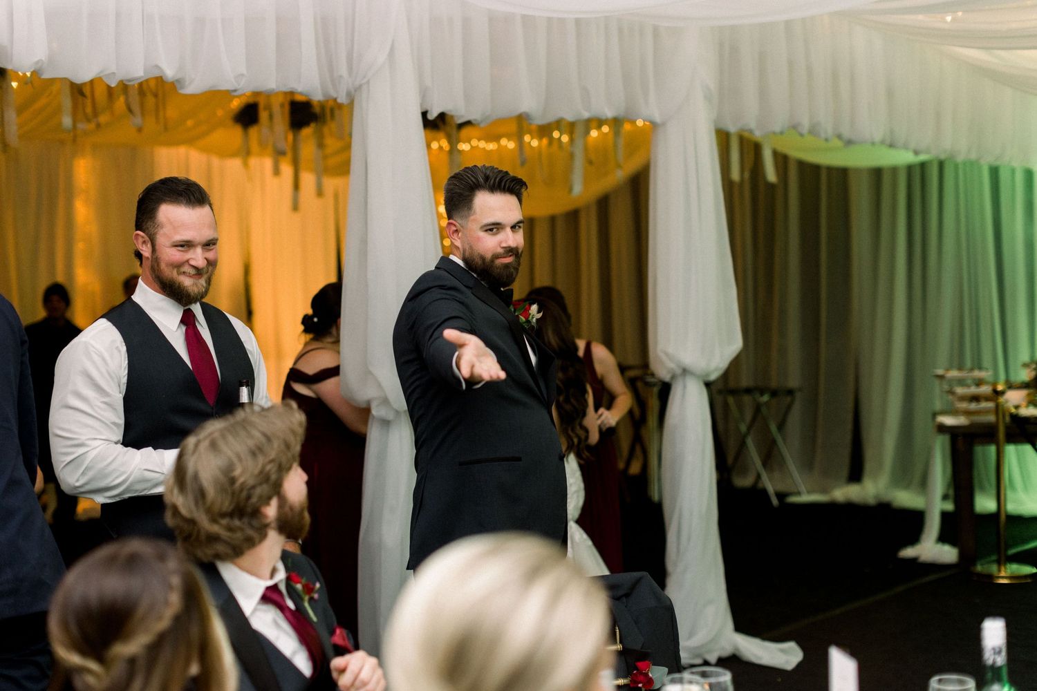 Wedding guests listen to speeches during reception with white draped backdrop.