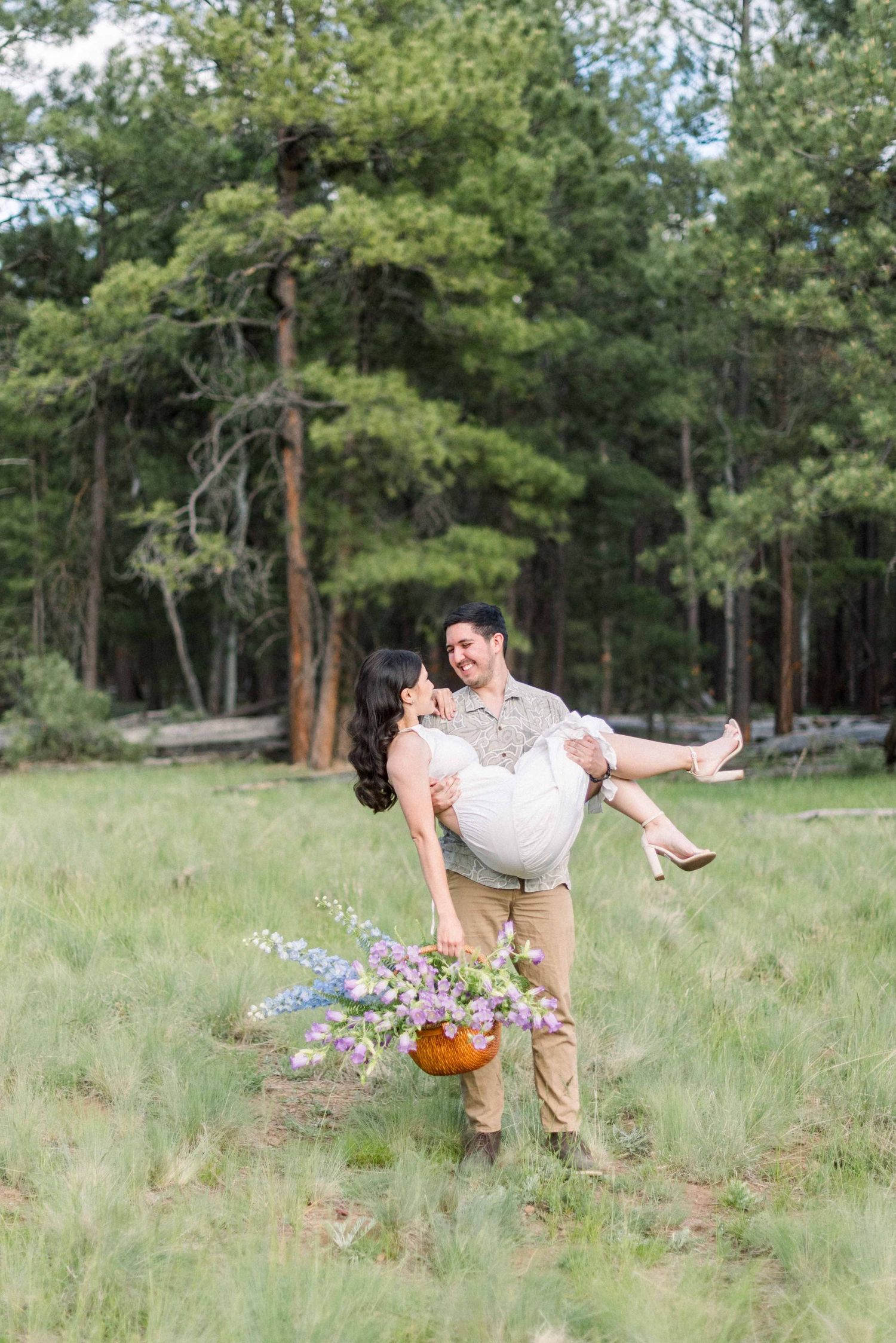 A romantic couple poses playfully in a meadow with pine trees as one lifts the other while holding a flower basket.