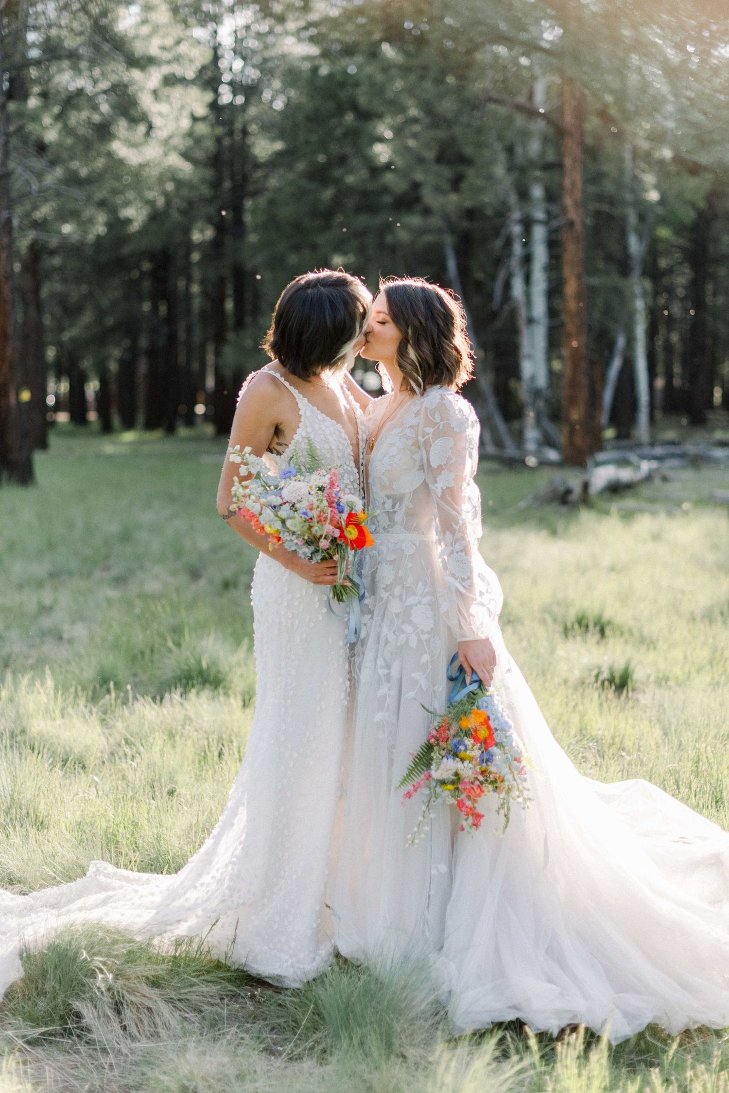 Two brides in white lace gowns share an intimate moment while holding colorful bouquets in a forest clearing.