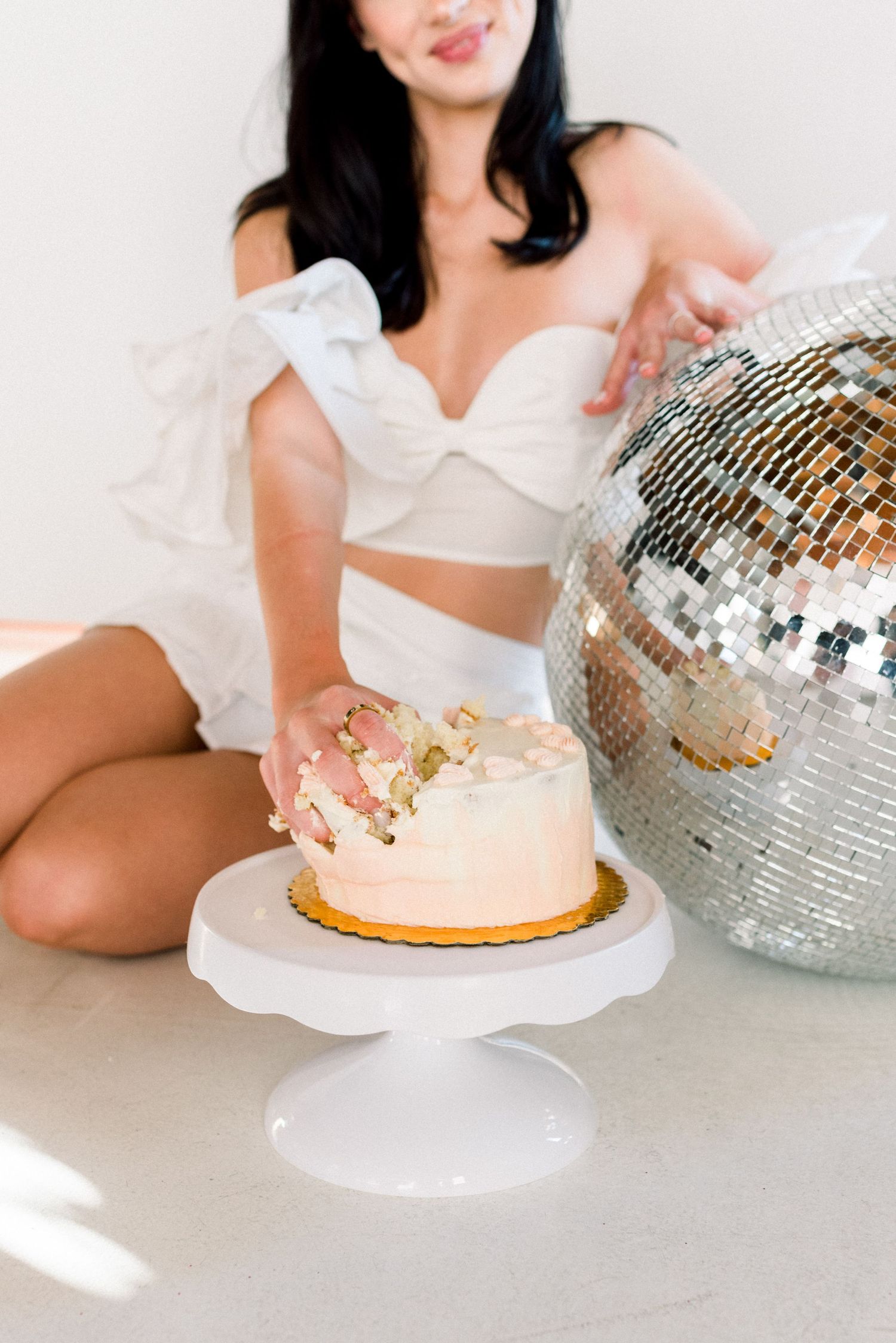 Person in white outfit enjoying cake beside large disco ball in bright setting.
