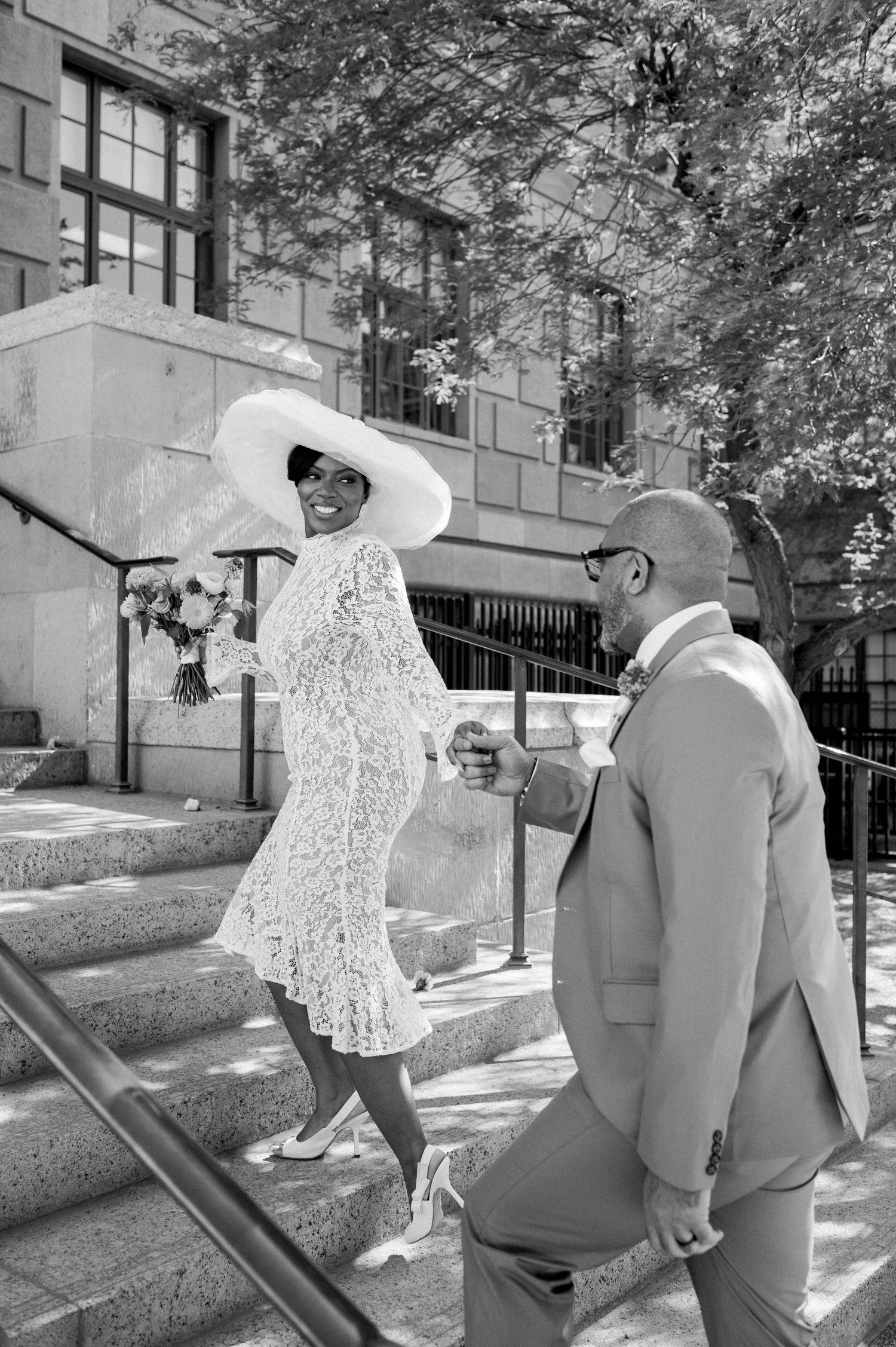 A couple ascends stone steps outside a building, with the person in white wearing a dramatic wide-brimmed hat.